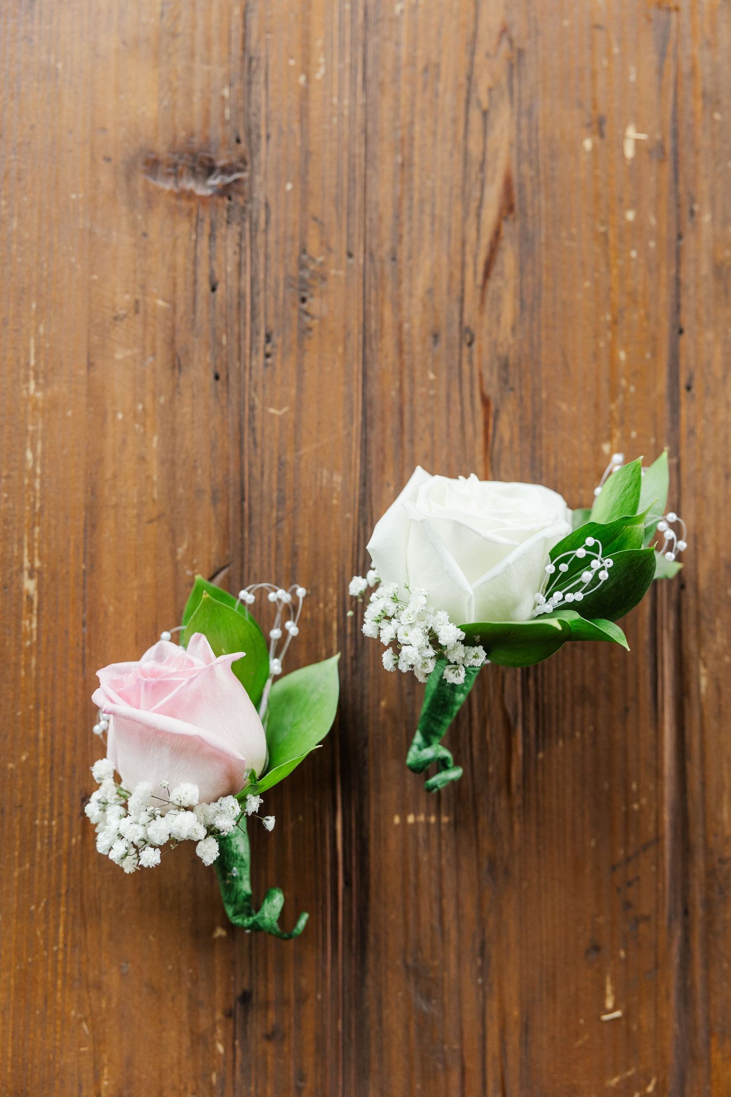 Two delicate wedding boutonnieres with pink and white roses and baby's breath on a wooden surface.