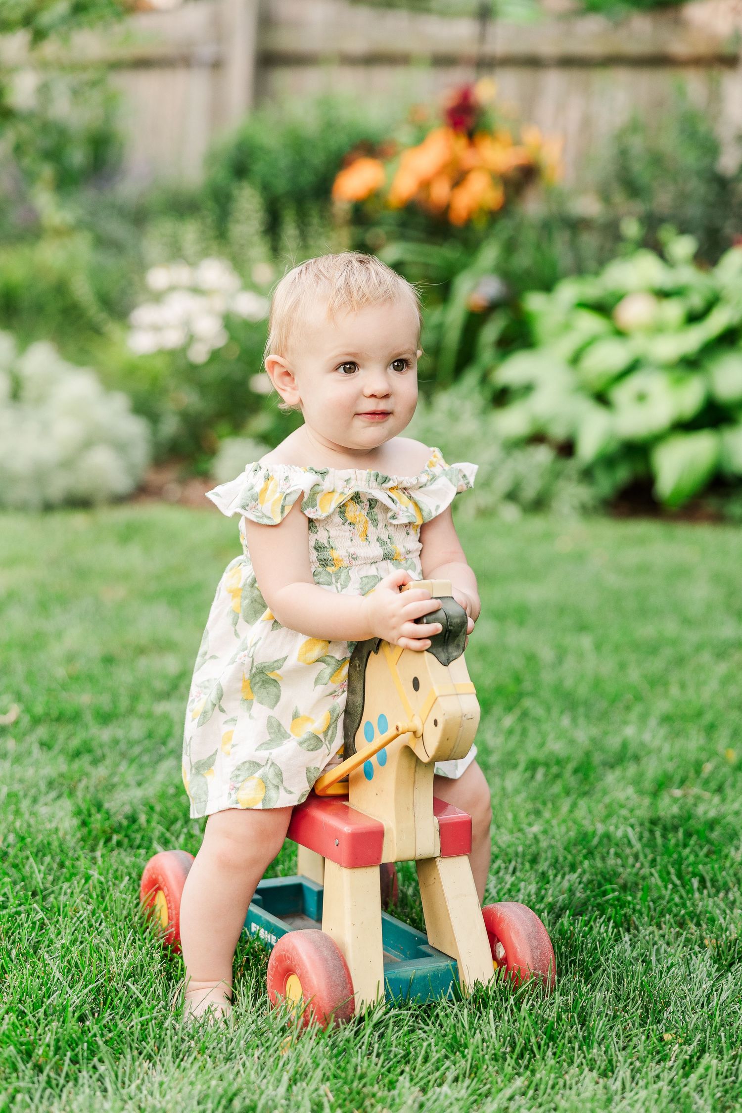 A small child in a ruffled floral dress plays on a wooden ride-on toy in a lush garden setting.