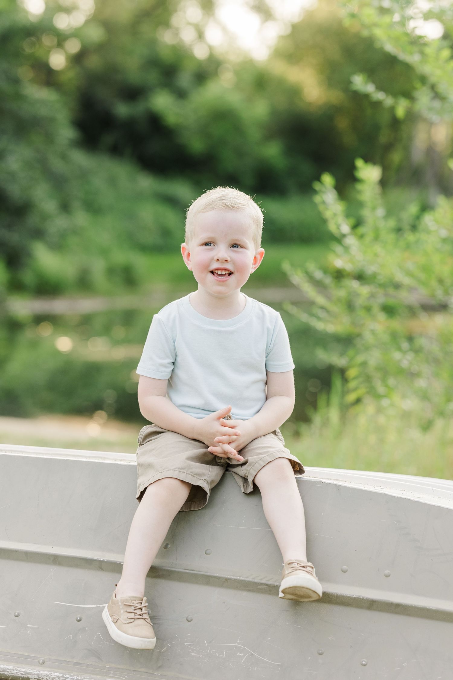 A young child in a white t-shirt and beige shorts sits on a concrete ledge with greenery in the background.