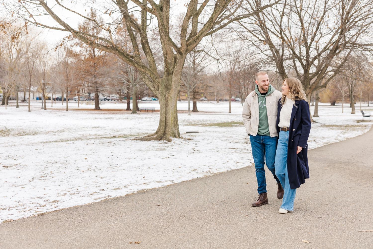 A couple takes a romantic winter stroll along a snowy path in the park with bare trees in the background.