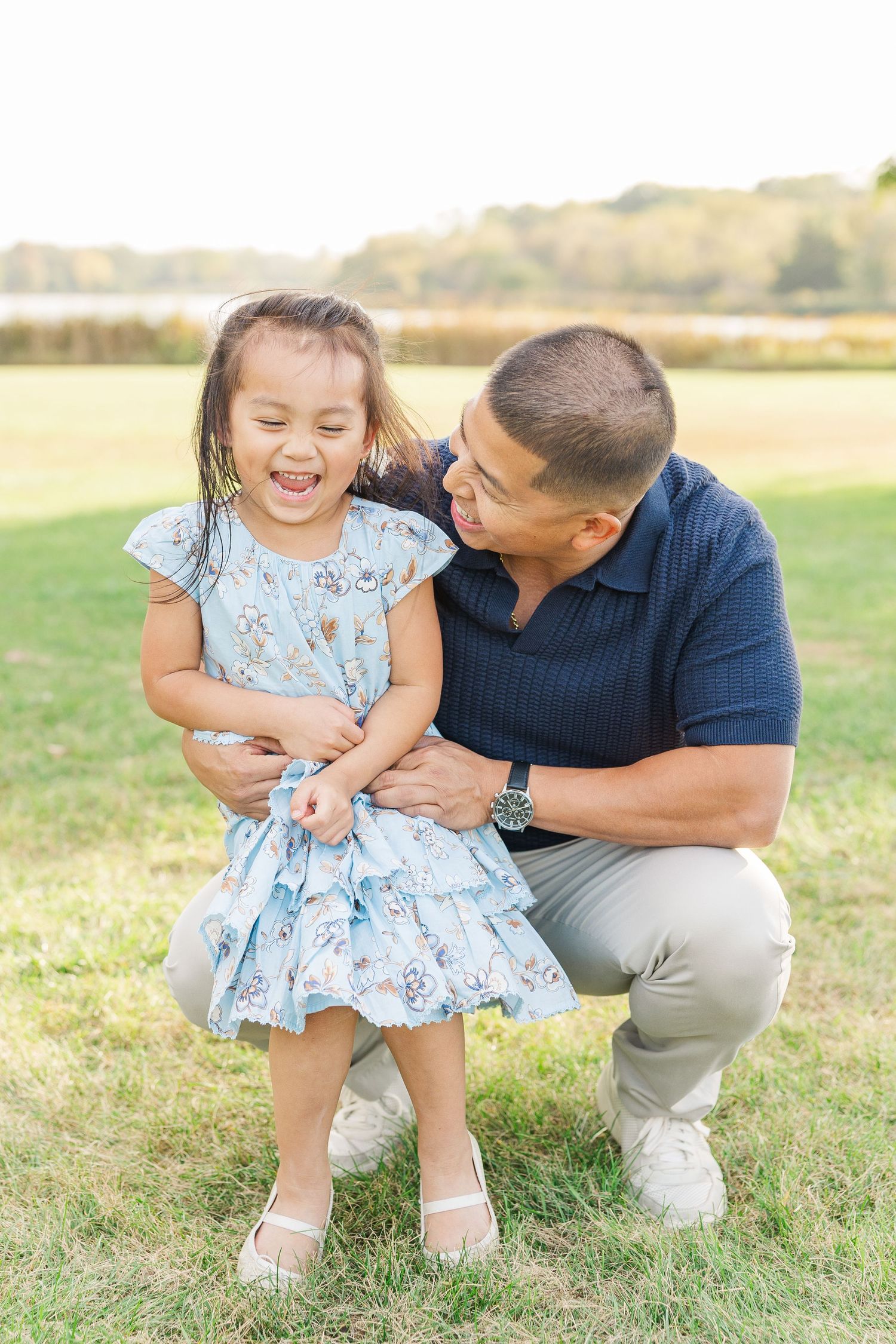A parent and child share a playful moment together in a sunny field while wearing coordinated blue and khaki outfits. Busse Woods