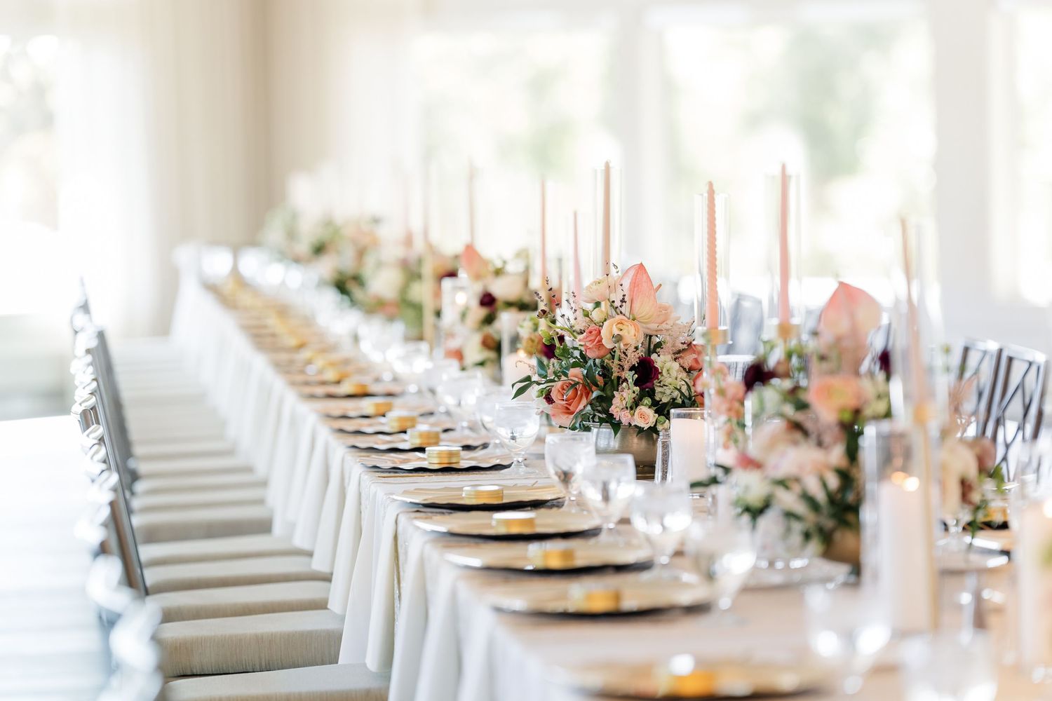 Long elegant wedding reception table with white linens, gold chargers, tall candles and blush floral centerpieces in a bright, airy venue.