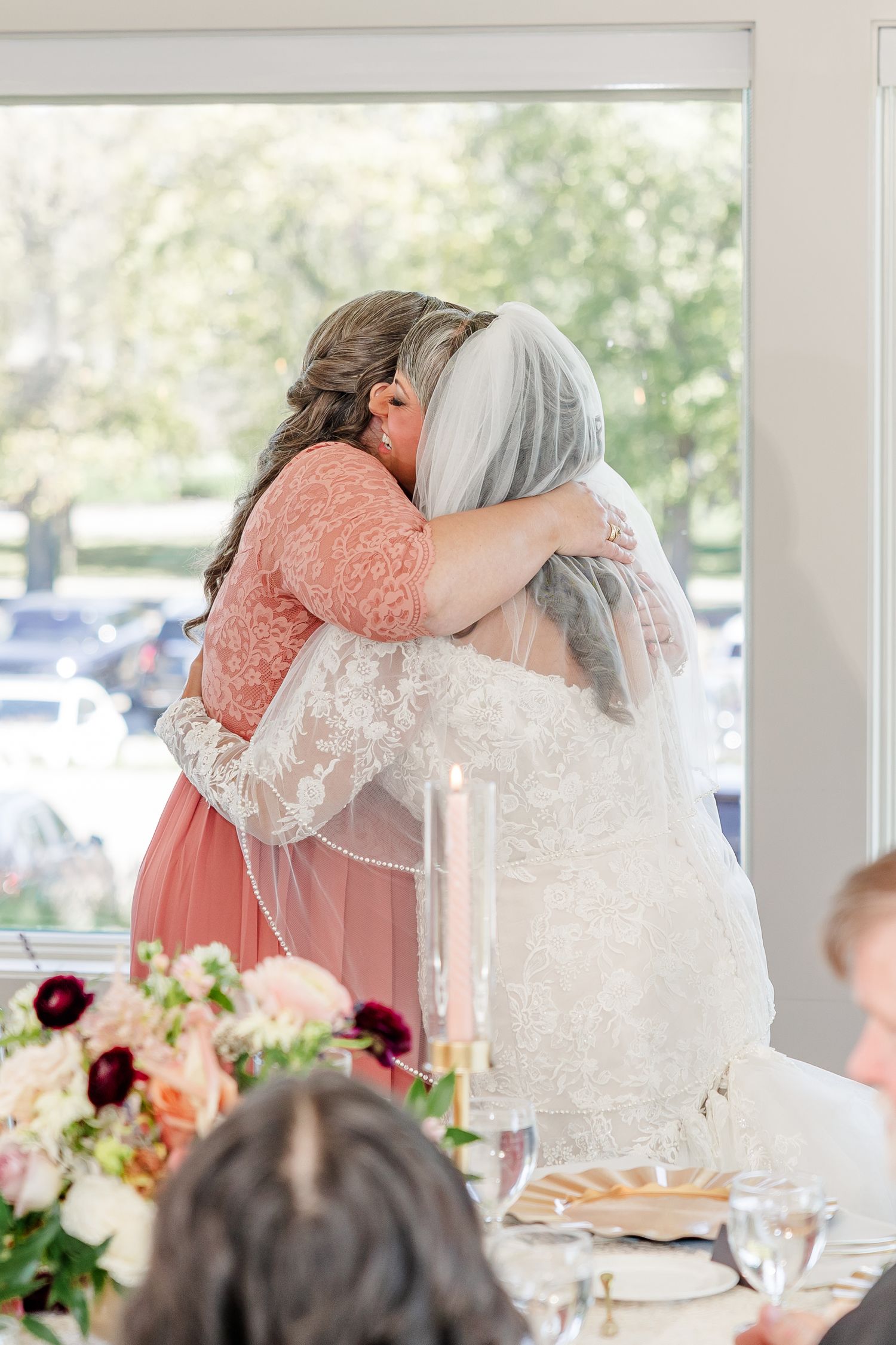 A bride in a lace gown and veil shares a warm hug with a woman in a coral lace dress beside a floral centerpiece at a reception.