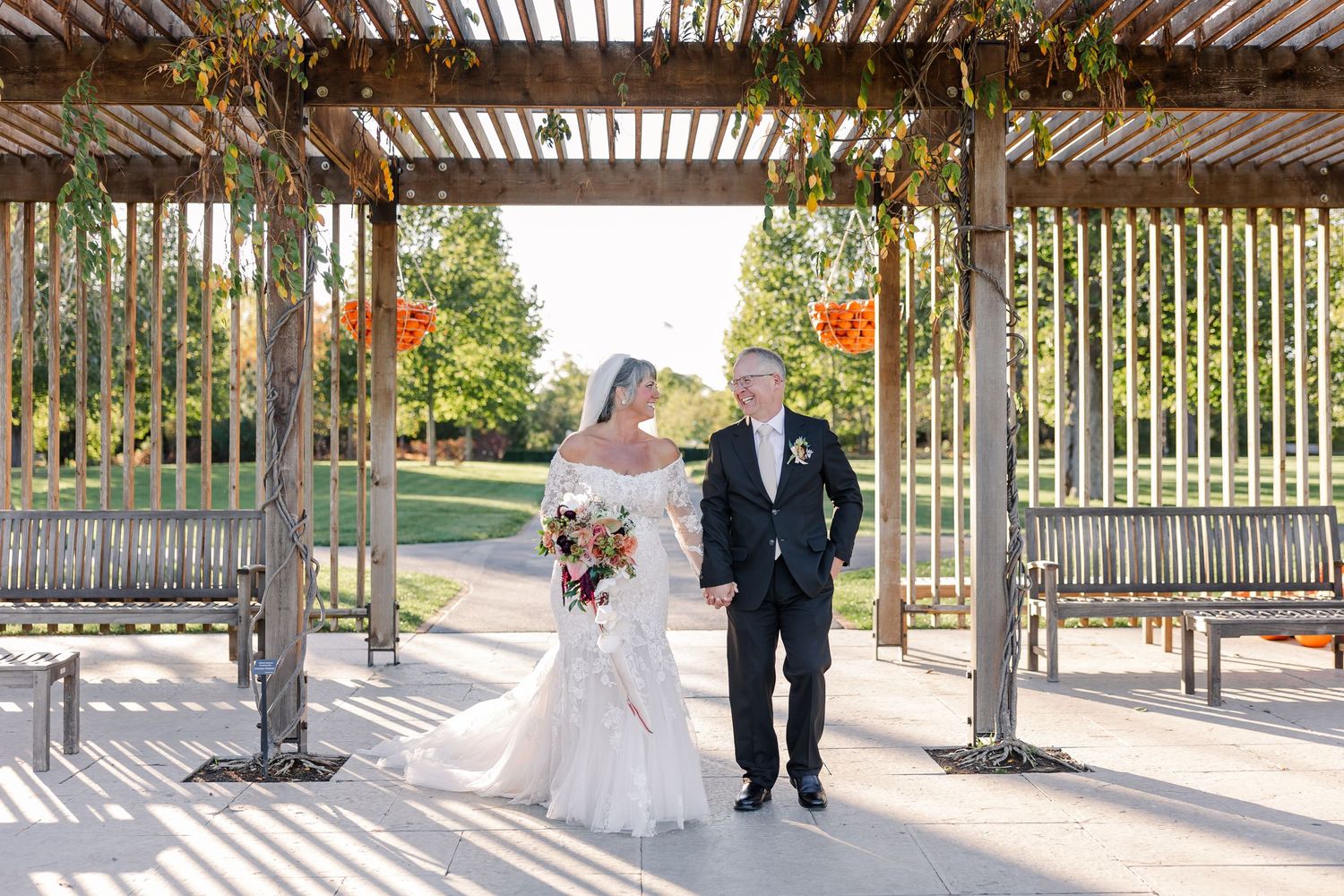Bride in a white lace gown holding a colorful bouquet walks hand in hand with a groom in a dark suit under a sunlit wooden pergola.