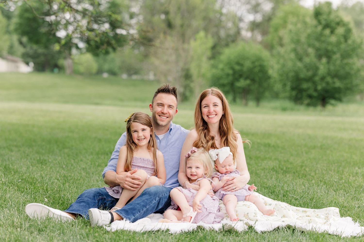 Happy family of four sitting on a blanket in a lush green park, parents smiling with two young daughters.