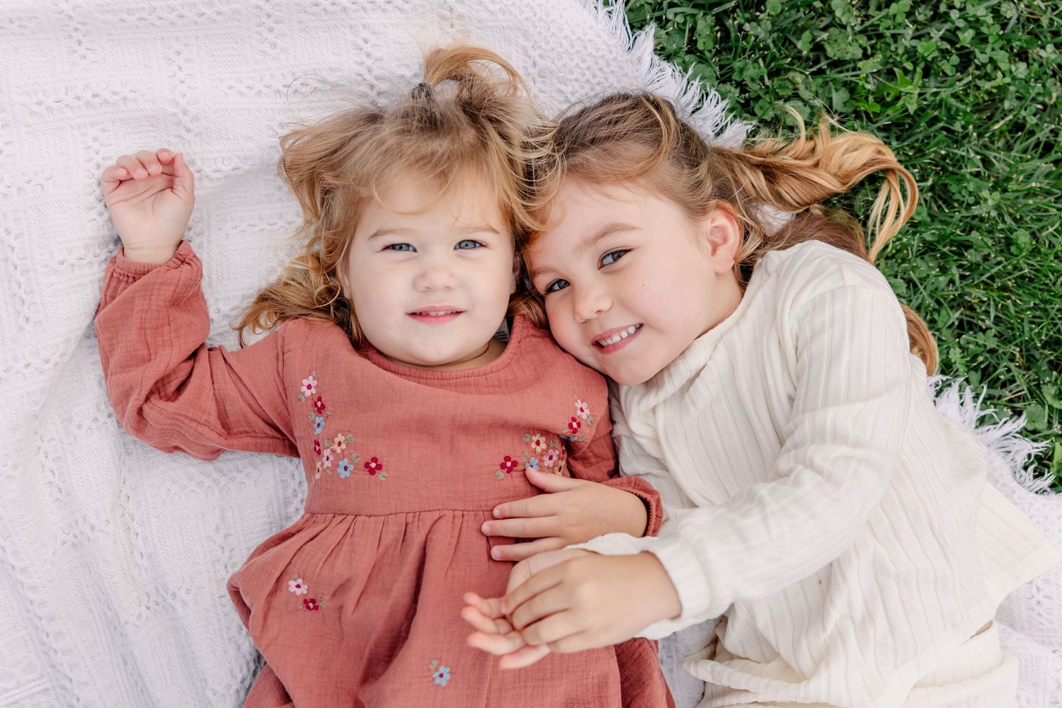 Two adorable little sisters lying on a blanket in the grass, smiling and hugging each other.