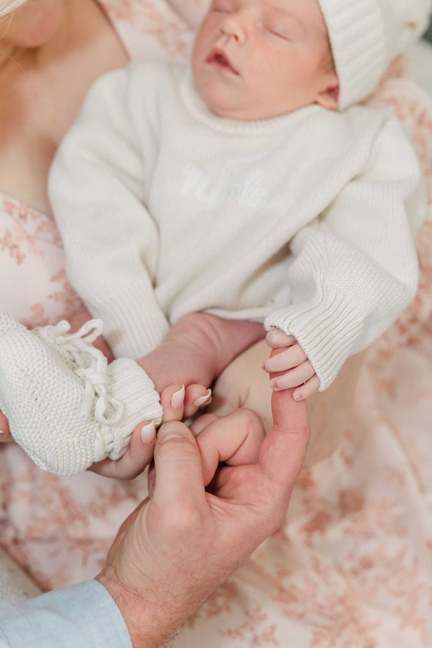Newborn baby in white knit outfit held by parents, tiny hand grasping adult finger in tender moment.