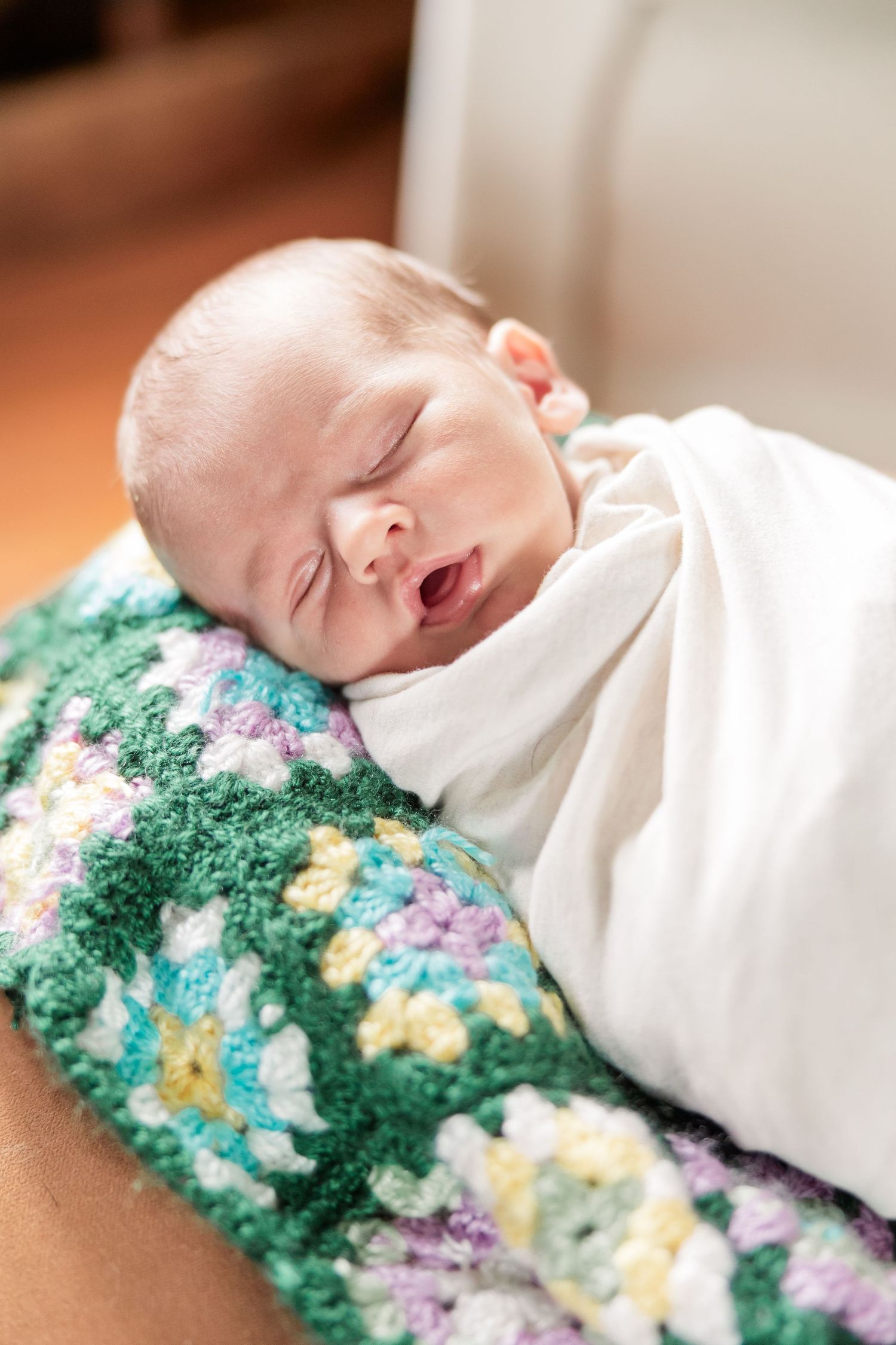 Peaceful newborn baby yawning while swaddled in white wrap on colorful crocheted granny square blanket.