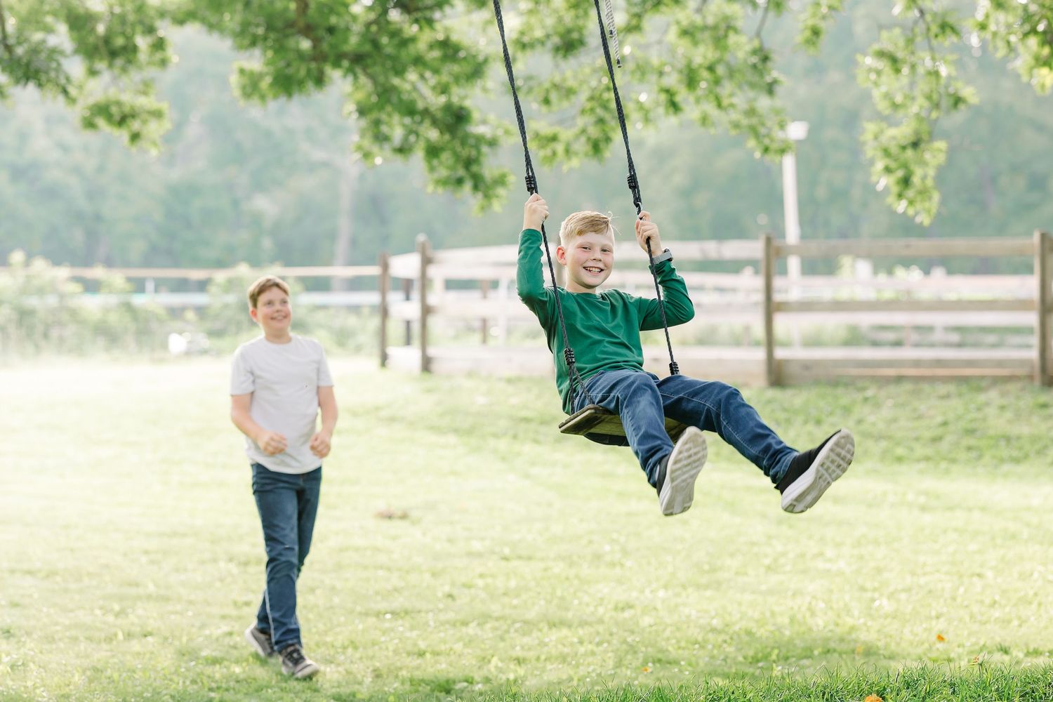 Young boy laughing while swinging on a rope swing in a sunny green park with an adult nearby.
