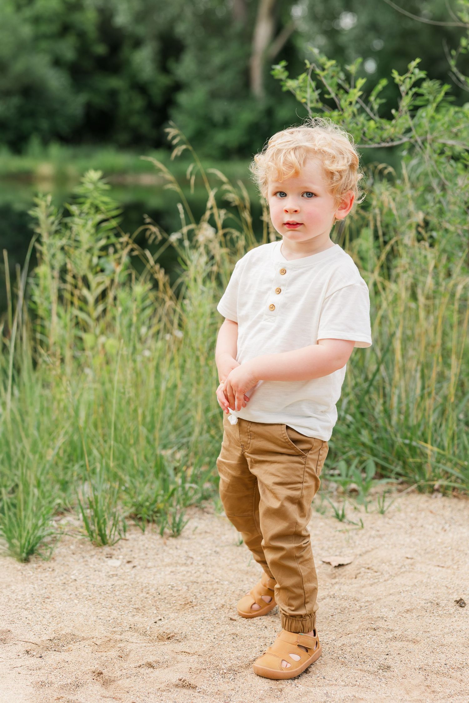 Adorable curly blonde toddler boy in white shirt and tan pants standing on sandy path near tall grass.