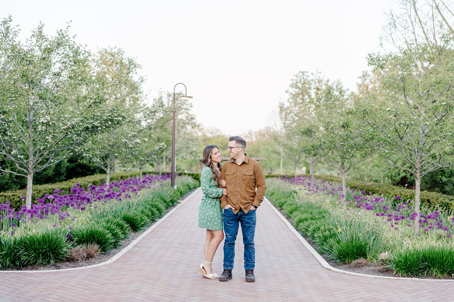 Couple shares a romantic moment on a garden path lined with purple flowers and green trees during an engagement session.