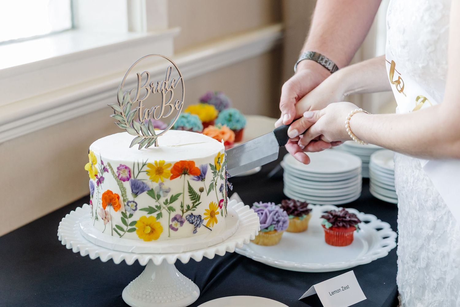 White wedding cake decorated with pressed flowers on a pedestal stand next to floral cupcakes being served.
