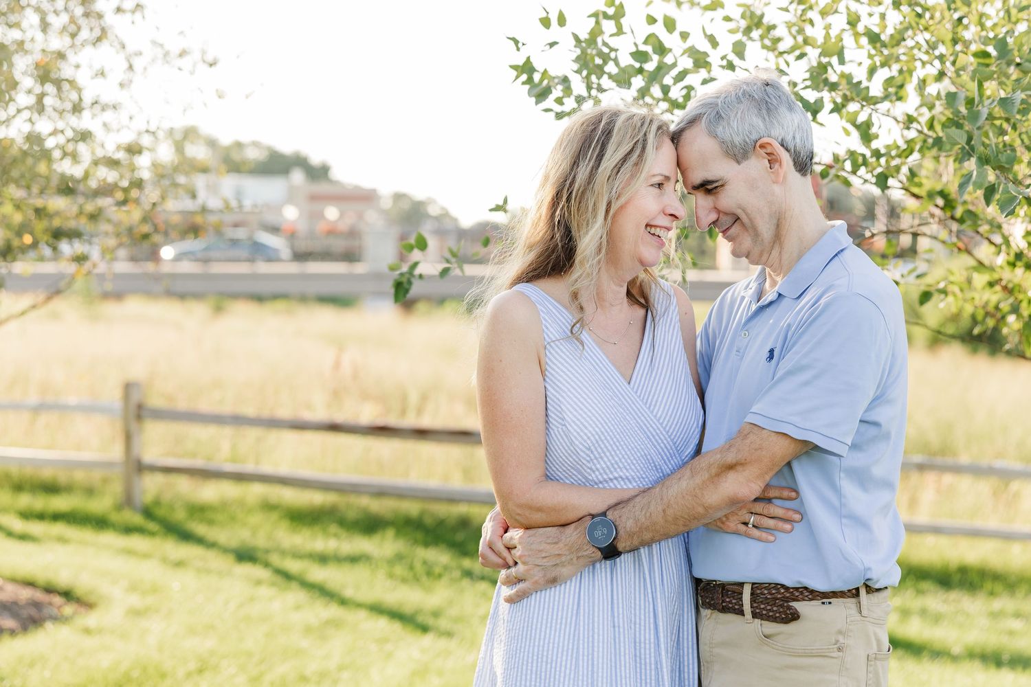 A smiling middle-aged couple embraces outdoors near a tree, with a sunny meadow and wooden fence in the background.