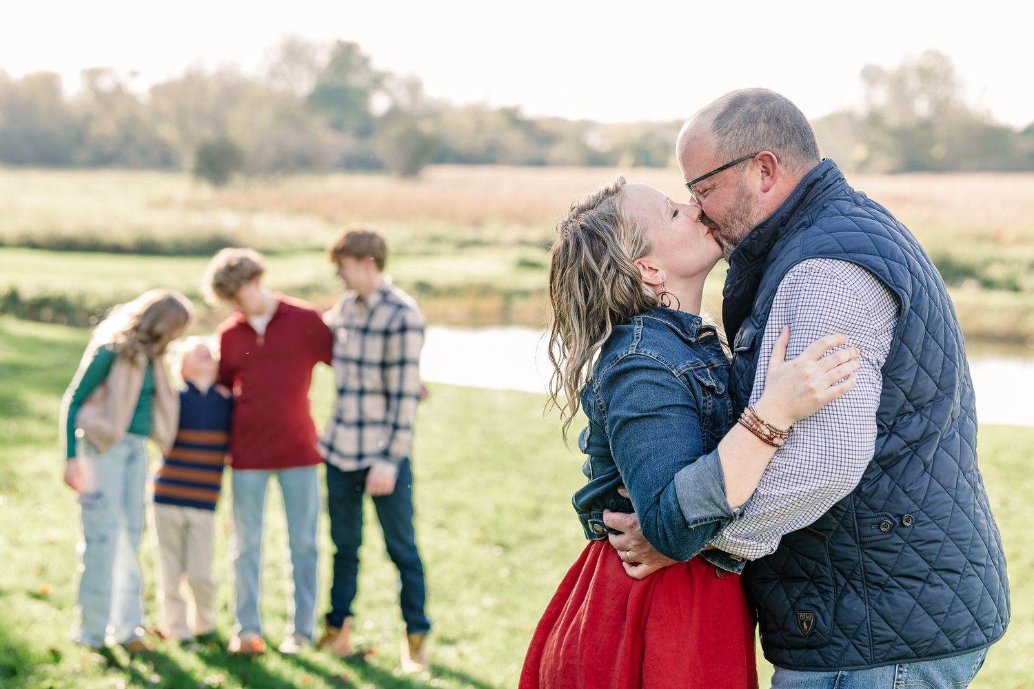 A man in a navy vest kisses a woman in a red skirt in a grassy field while four children stand together in the background.