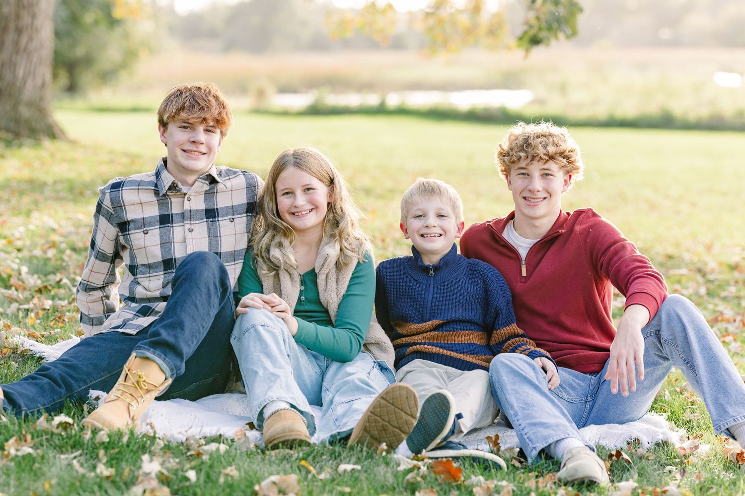 Four smiling siblings in plaid, green, navy and red sweaters sit close together on a blanket in a sunny autumn park with fallen leaves.
