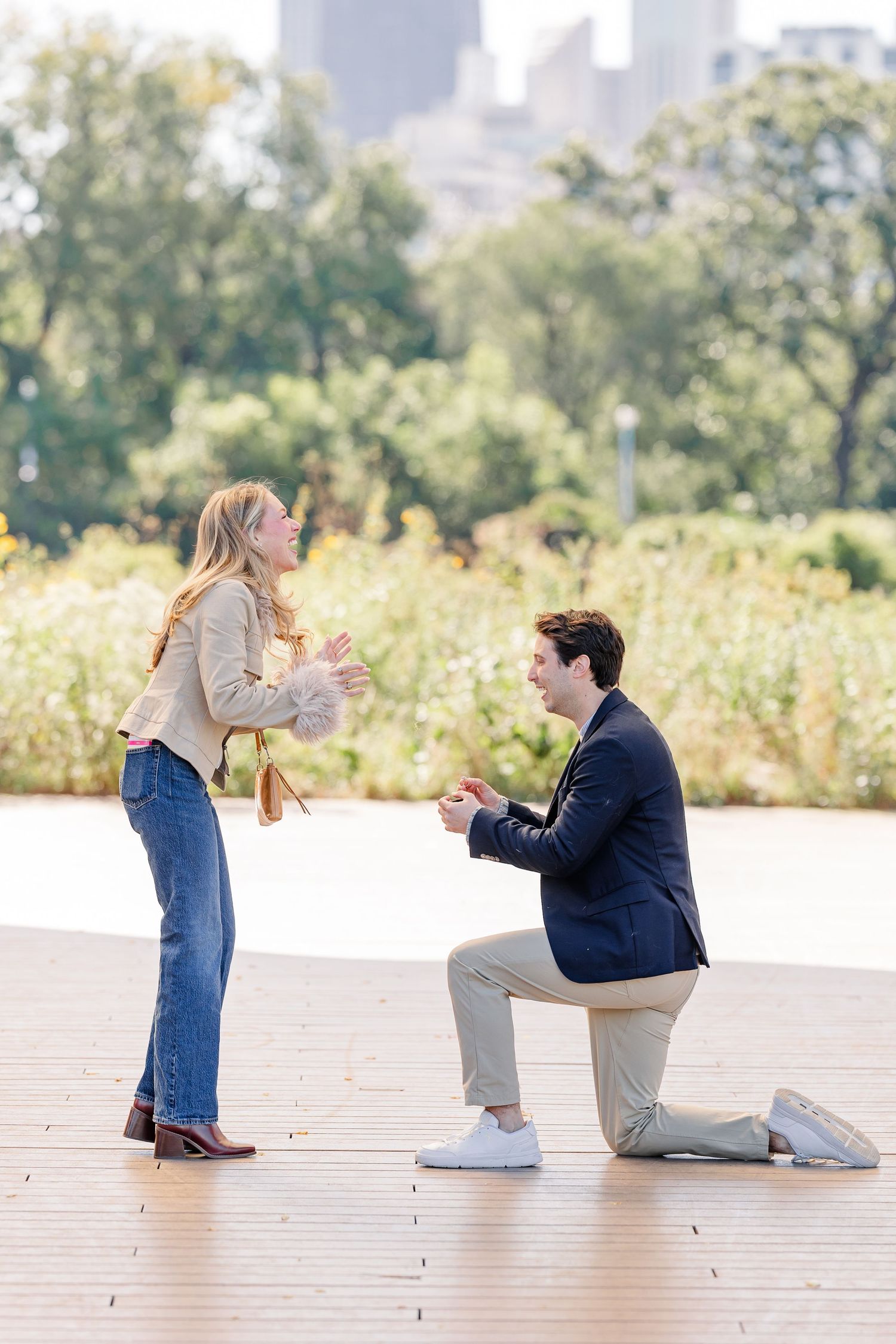 Man proposes on one knee to surprised woman on wooden deck in park with city skyline backdrop.