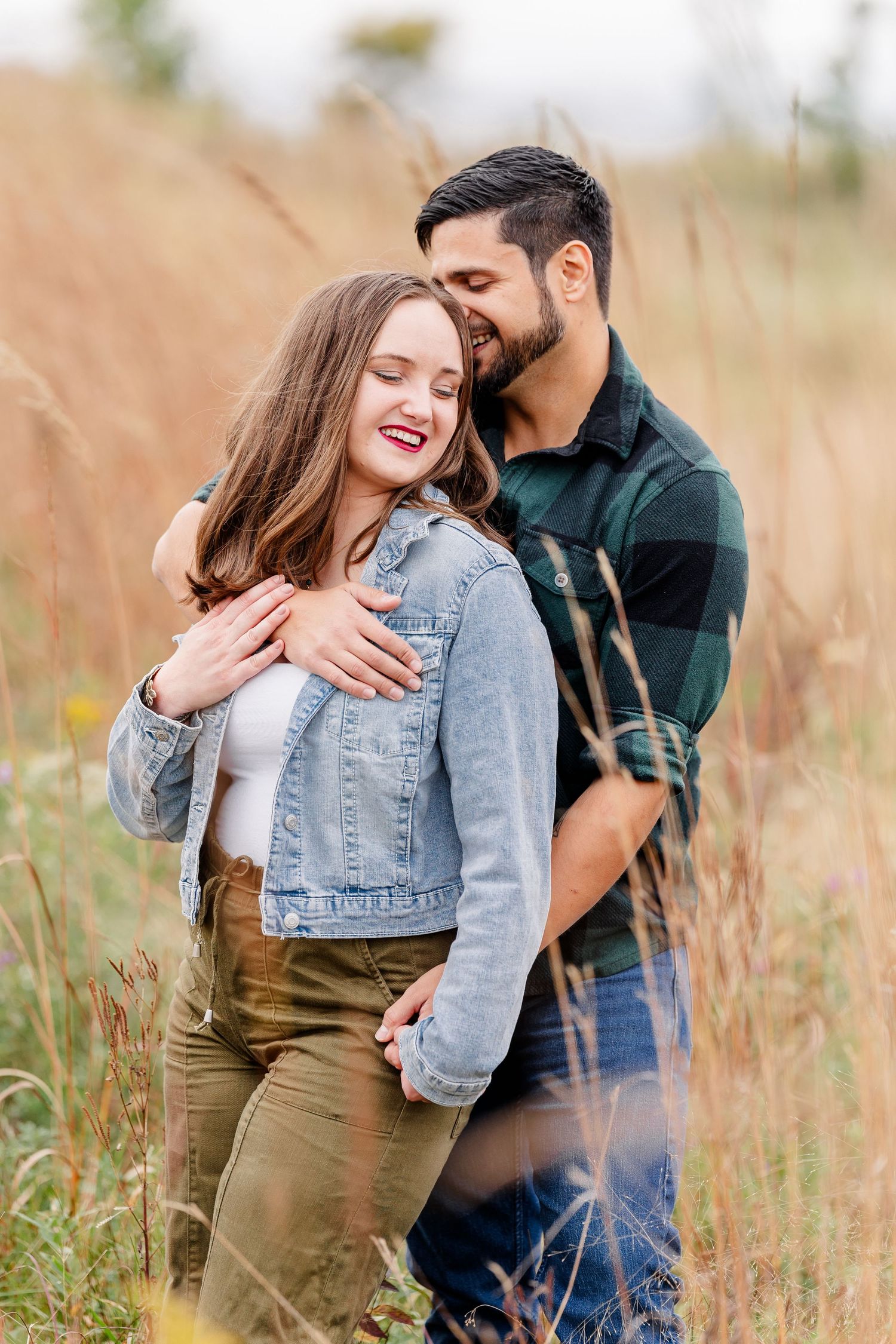 Close-up of a joyful couple cuddling in tall prairie grass, she wears a denim jacket and he wears a green plaid shirt.