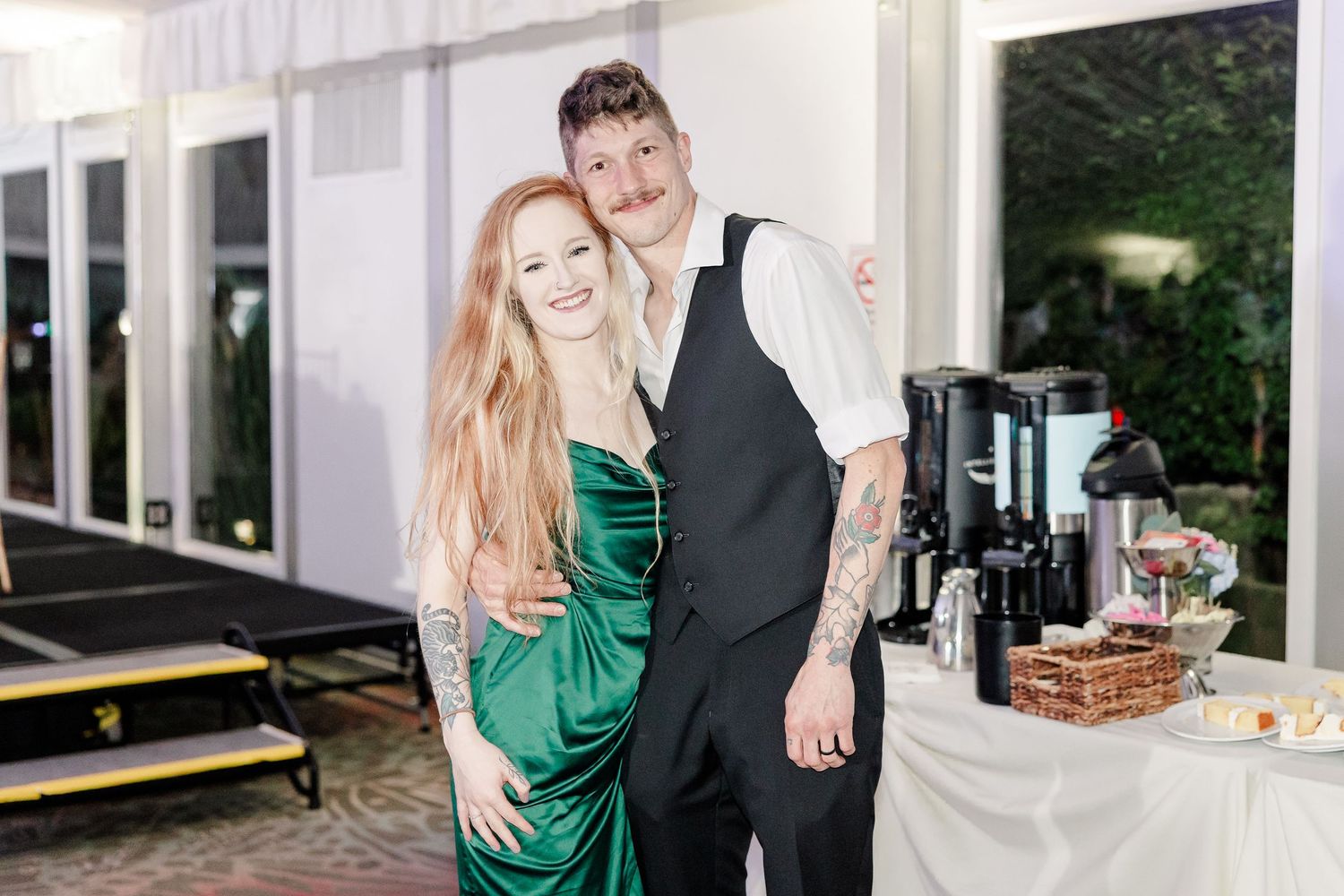 Woman in emerald green dress posing with tattooed man in vest at wedding reception.