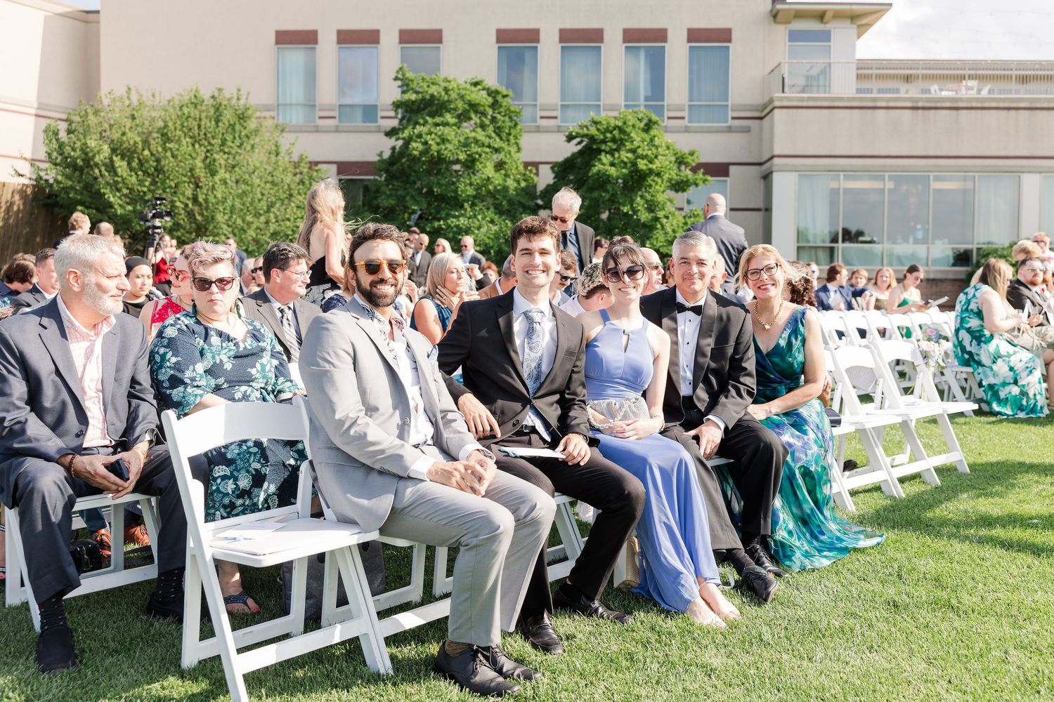 Wedding guests seated in white chairs outdoors smile during sunny garden ceremony.