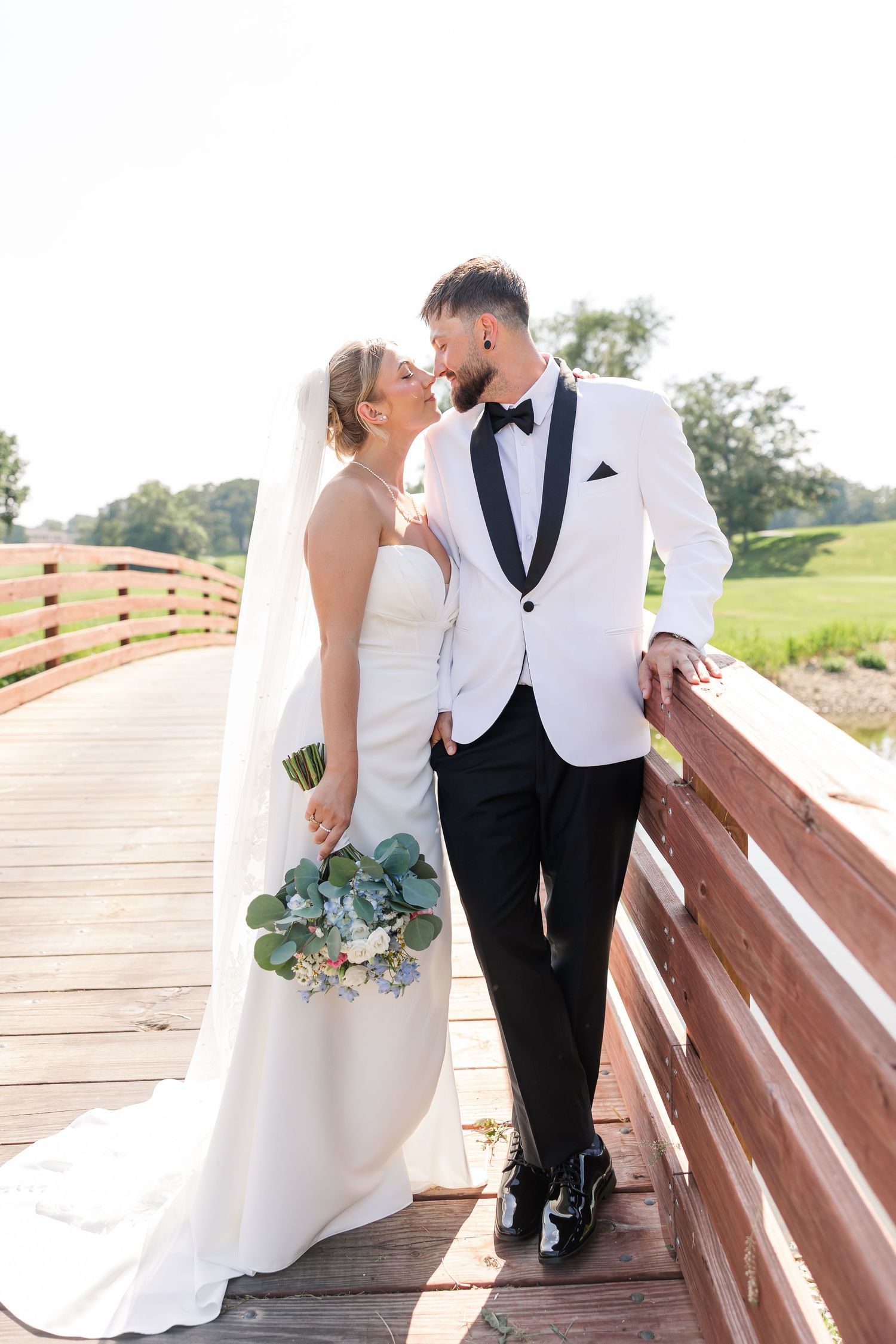 Bride and groom sharing romantic moment on wooden bridge, bride holding eucalyptus bouquet in white gown.