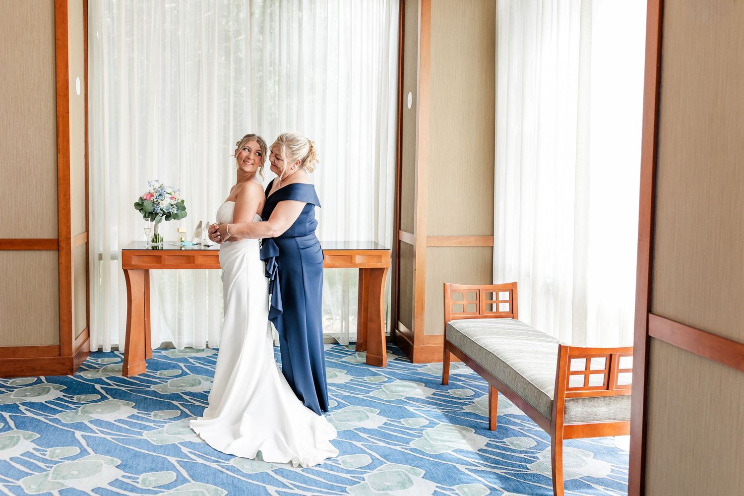 Bride in white gown shares emotional hug with woman in blue dress in elegant venue.