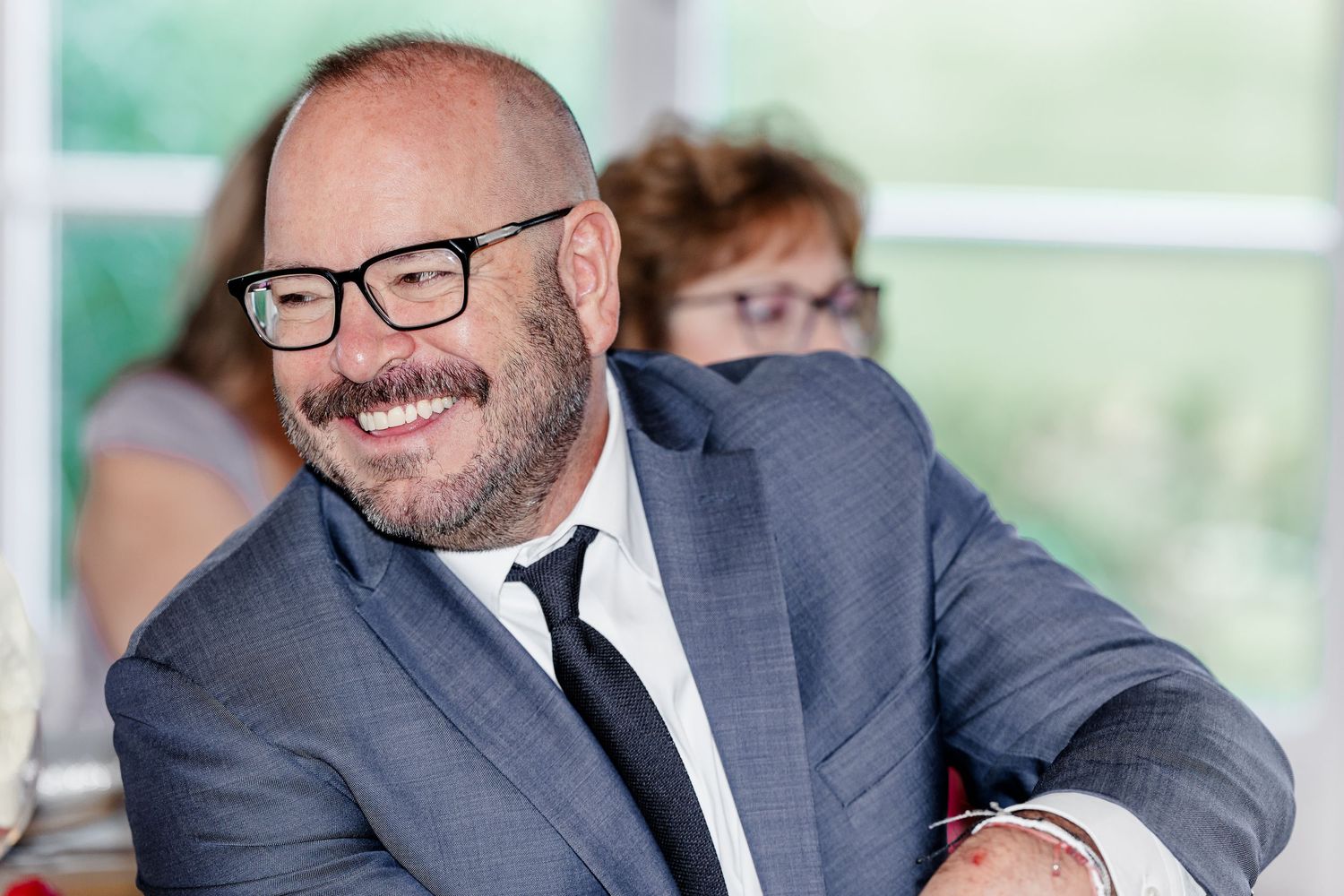 Smiling bald man with beard wearing gray suit and black tie at wedding reception event.