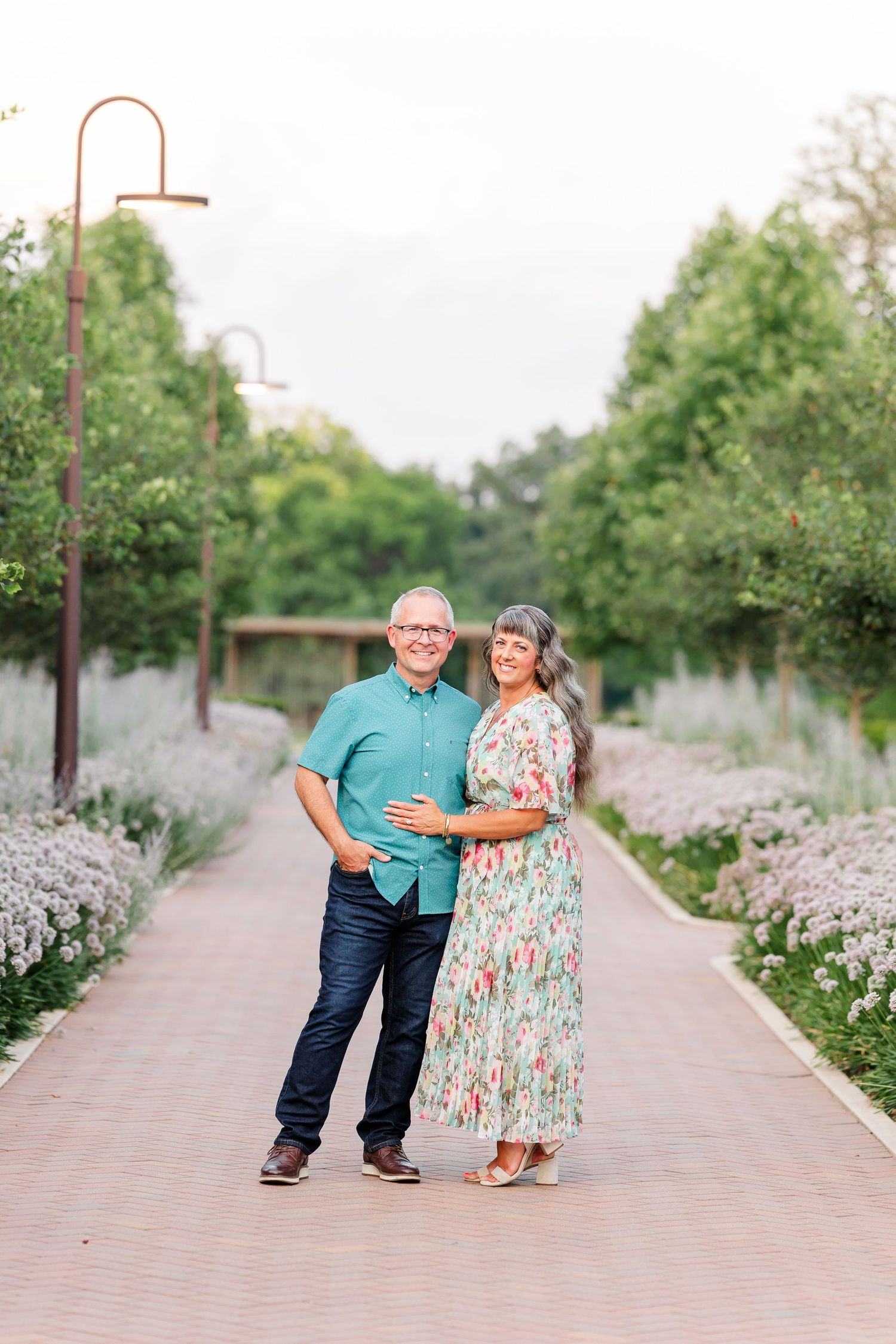 A smiling couple poses on a brick garden path lined with blooming flowers and lush green trees.