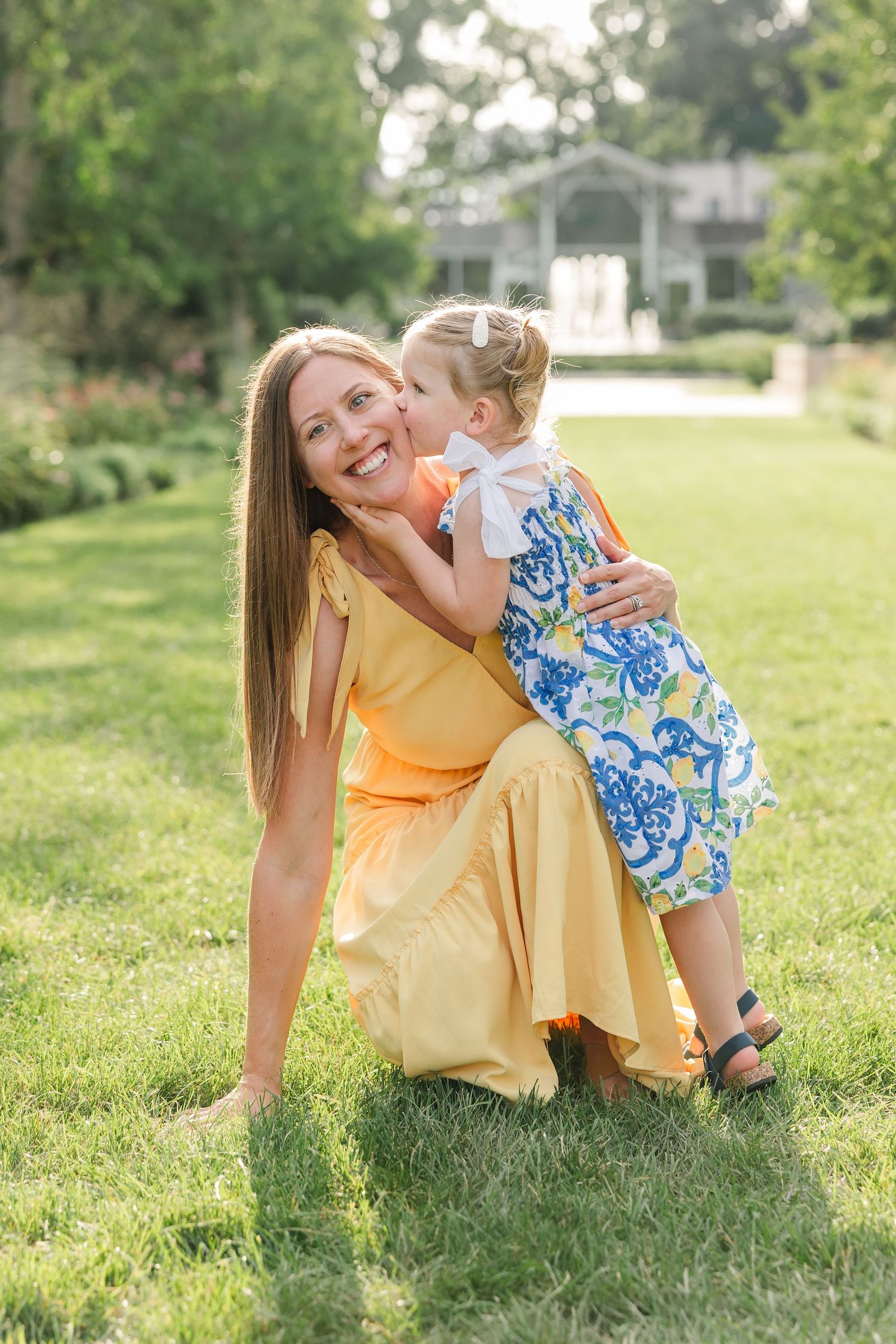 Joyful mother in yellow dress kneeling on grass as young daughter in floral outfit hugs her cheek.