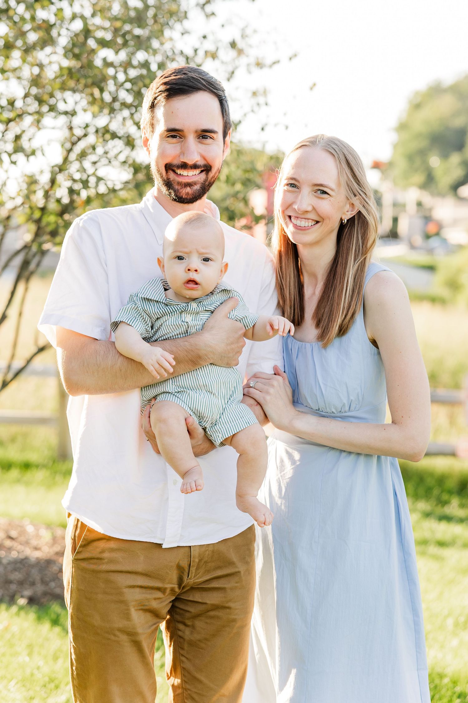 Smiling couple holding their baby infant outdoors in a sunny park setting.