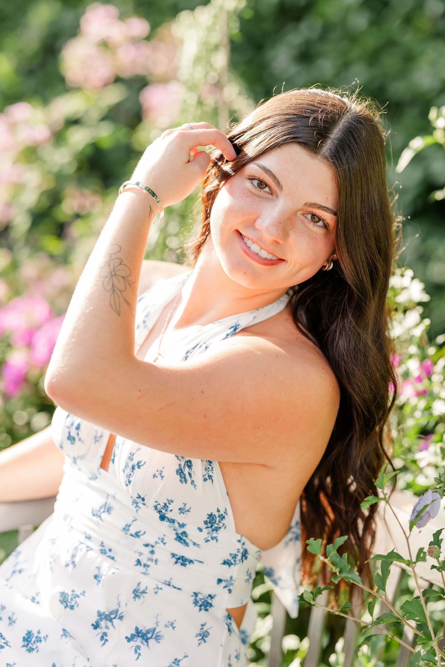 Young woman in floral dress smiling outdoors among pink blooming flowers on a sunny day.