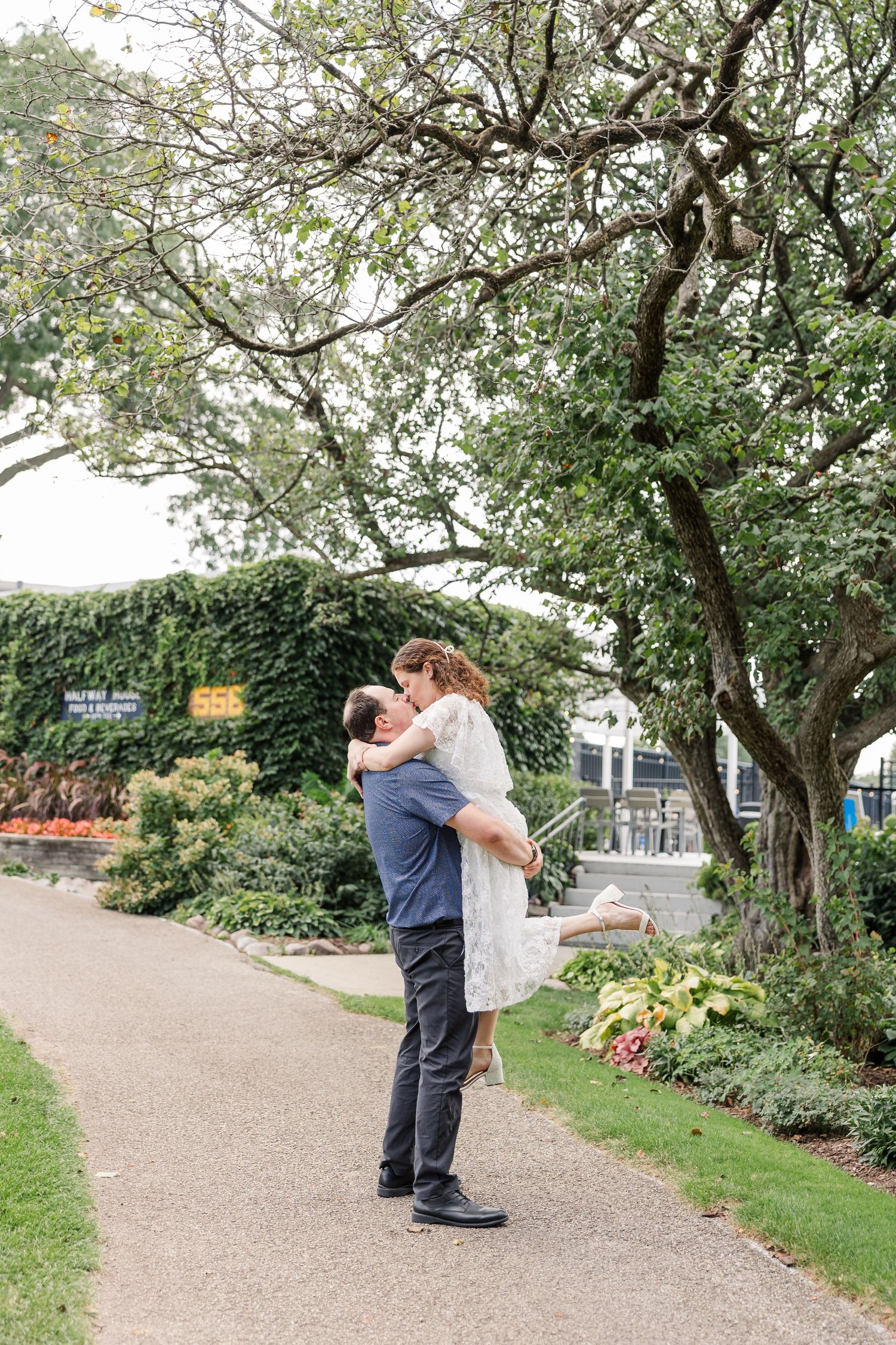 Groom lifting bride in white lace dress outdoors on garden pathway surrounded by lush greenery.