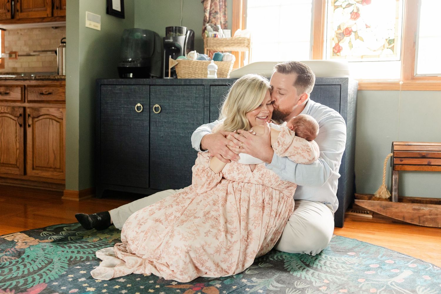 New parents sitting on floor sharing tender kiss over their sleeping newborn baby.