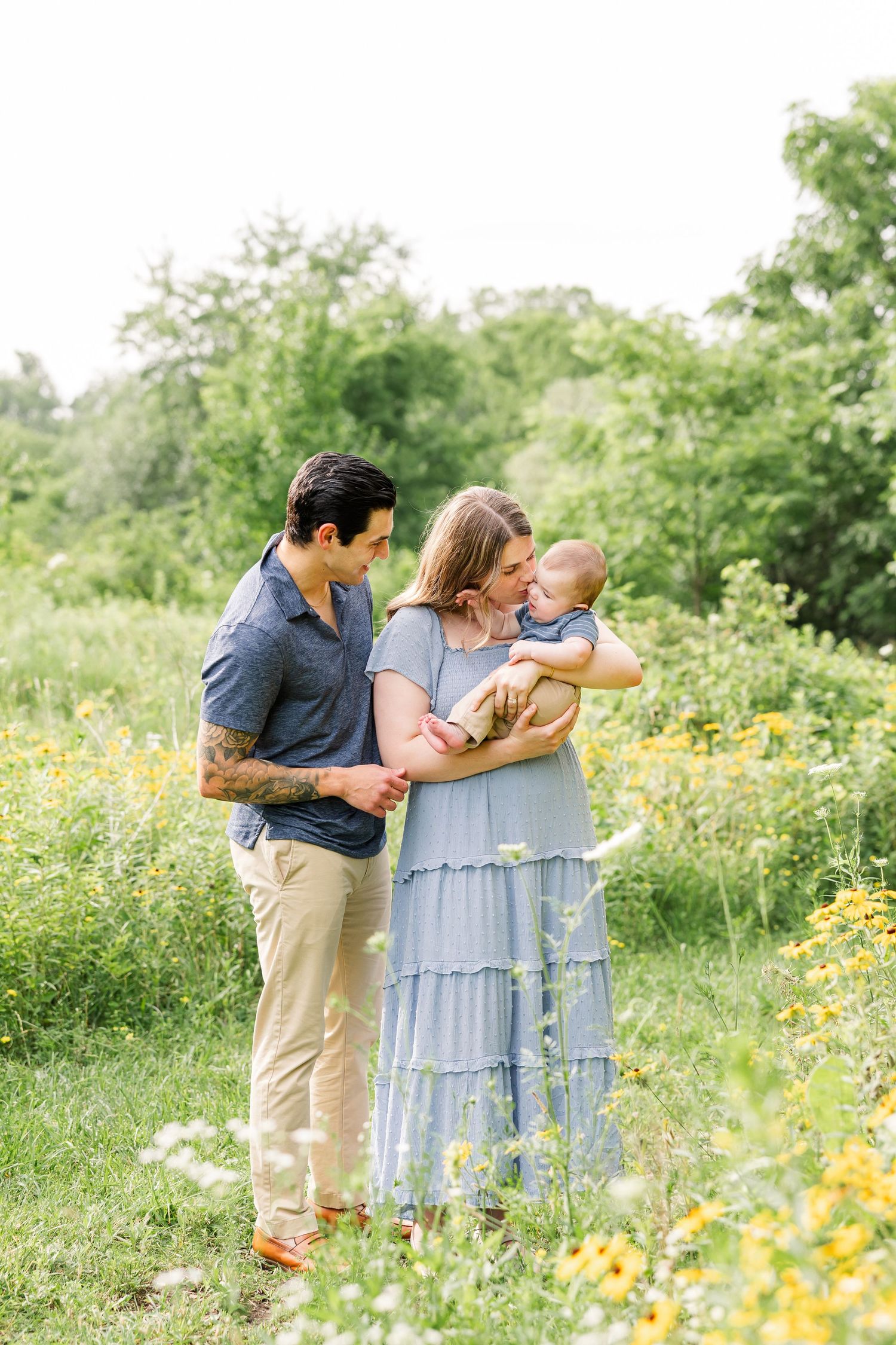 Young family with mother in blue dress and father with tattoos admiring baby in sunny wildflower meadow.