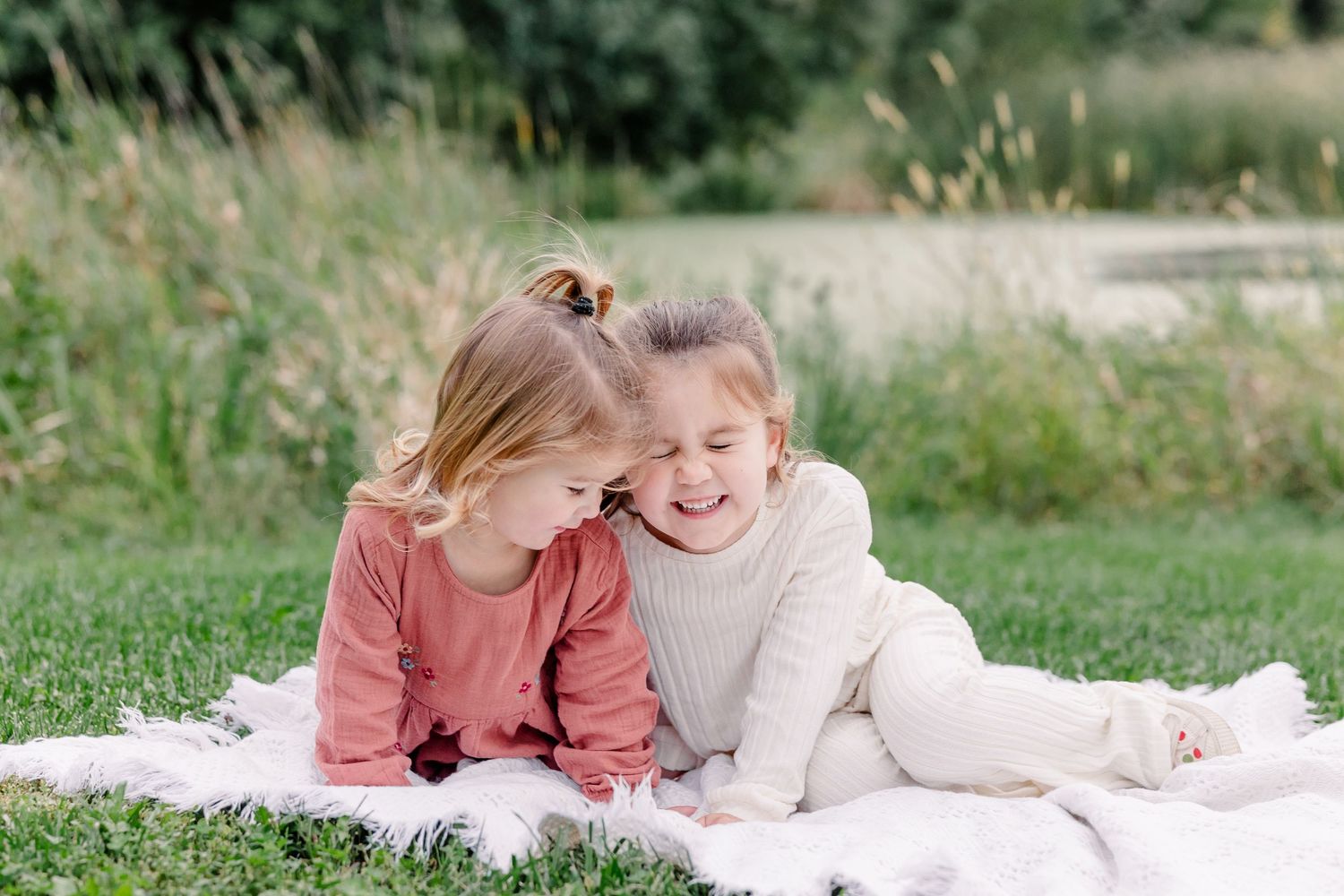 Two little girls in pink and cream outfits giggling together on a white blanket in a park.