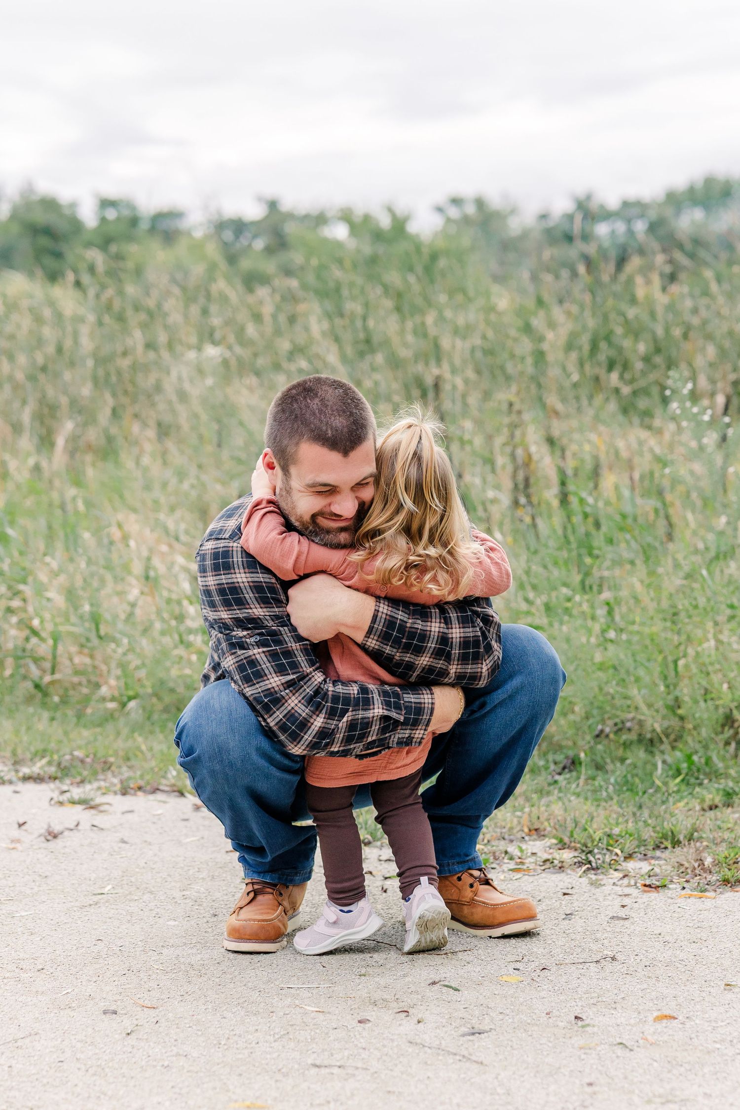 A father crouches down to embrace his young child on a path surrounded by tall green grass outdoors.
