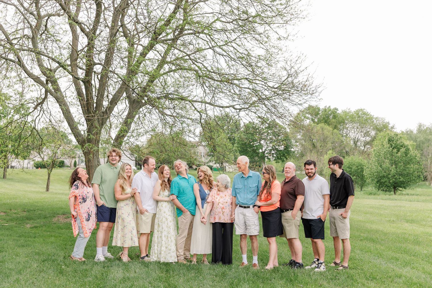 A large extended family group of about twelve people stands together smiling under a tree on a sunny green lawn.