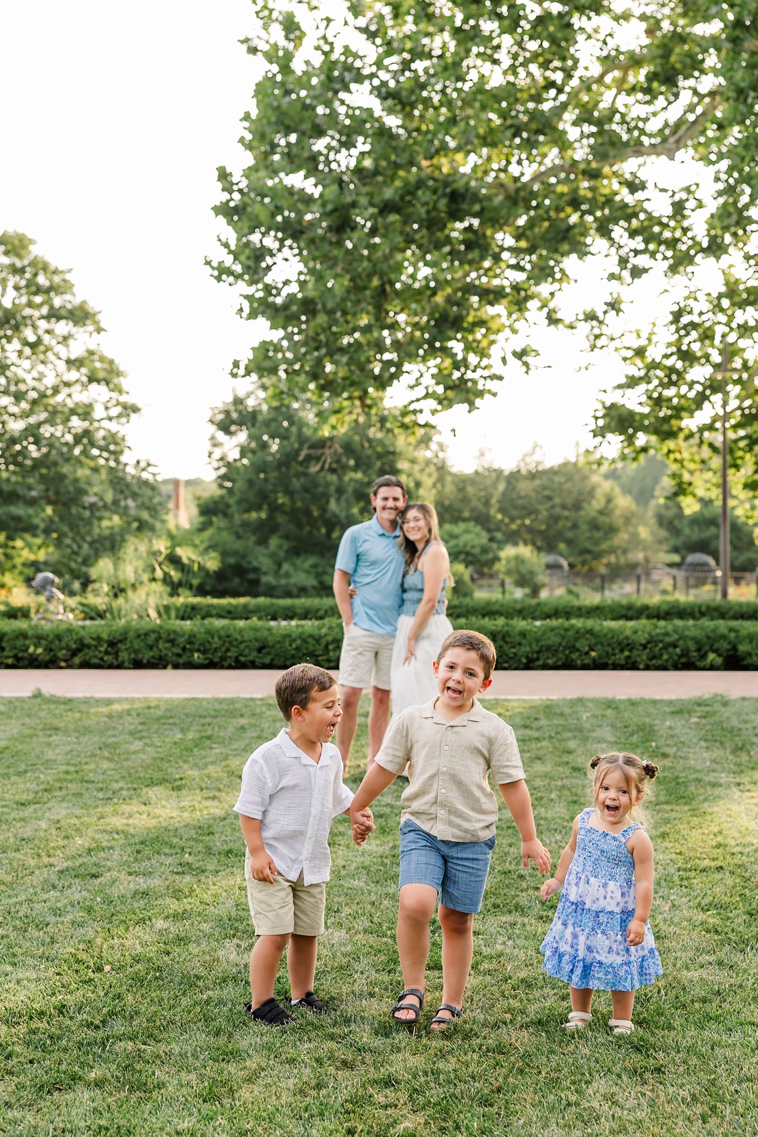 Happy family with three young children holding hands on green grass in a sunny park with lush trees.