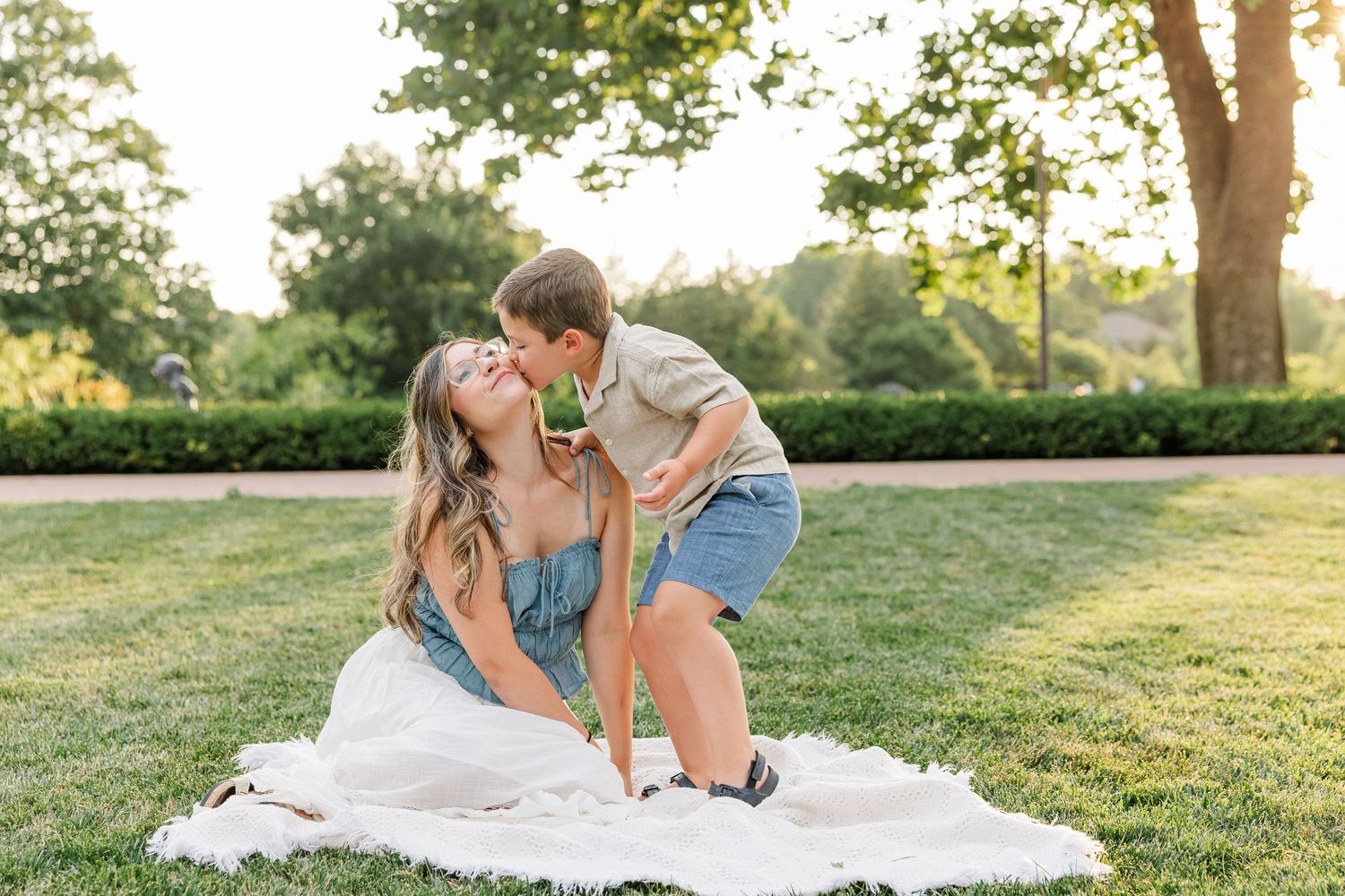 Mother sitting on white blanket in park receiving kiss from young boy in golden sunset light.