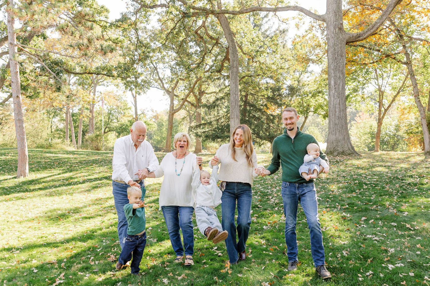Multi-generational family walking and laughing together in a lush green park during autumn family photo session.