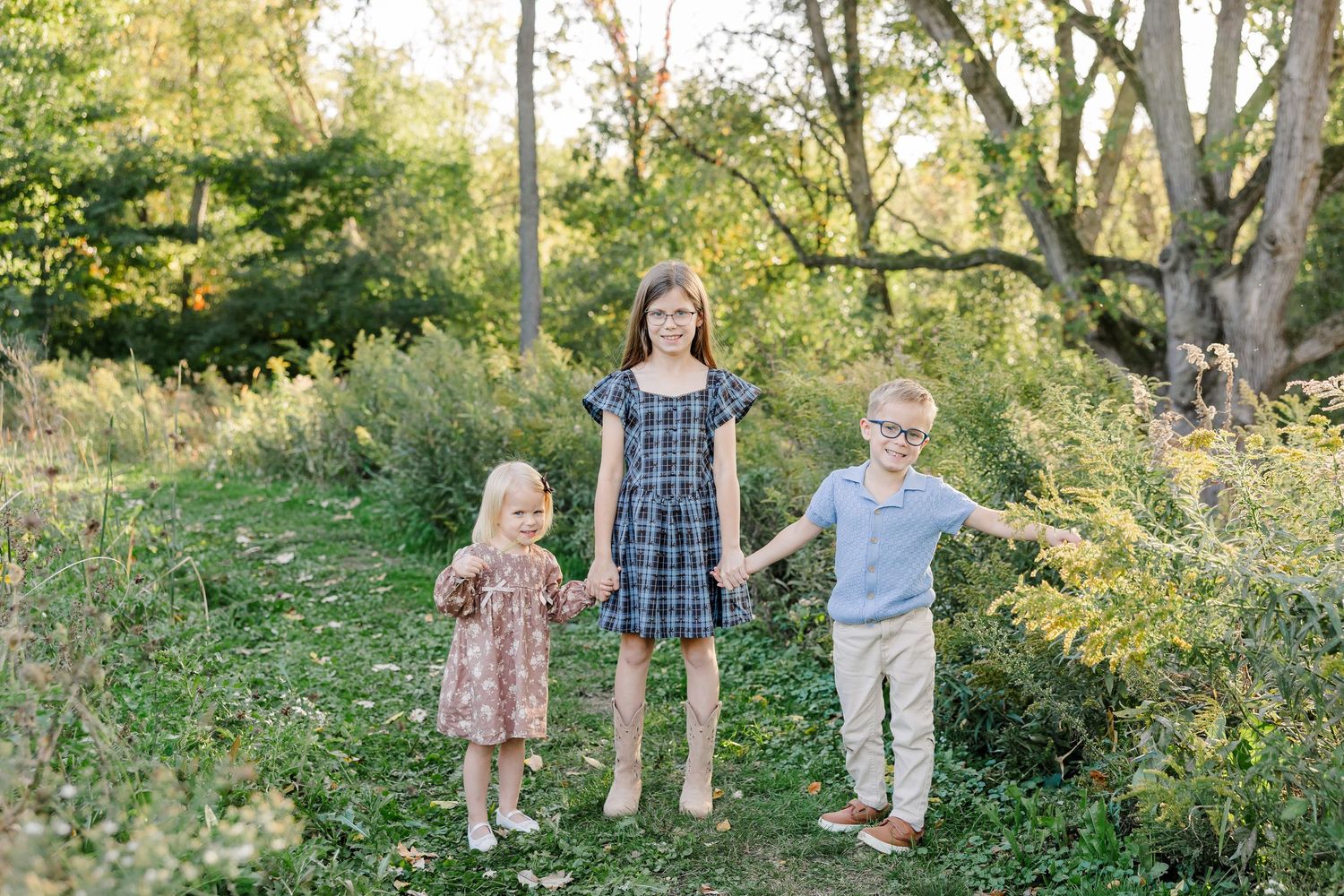 Two sisters and their brother holding hands in a sunlit backyard in St. Charles, IL.