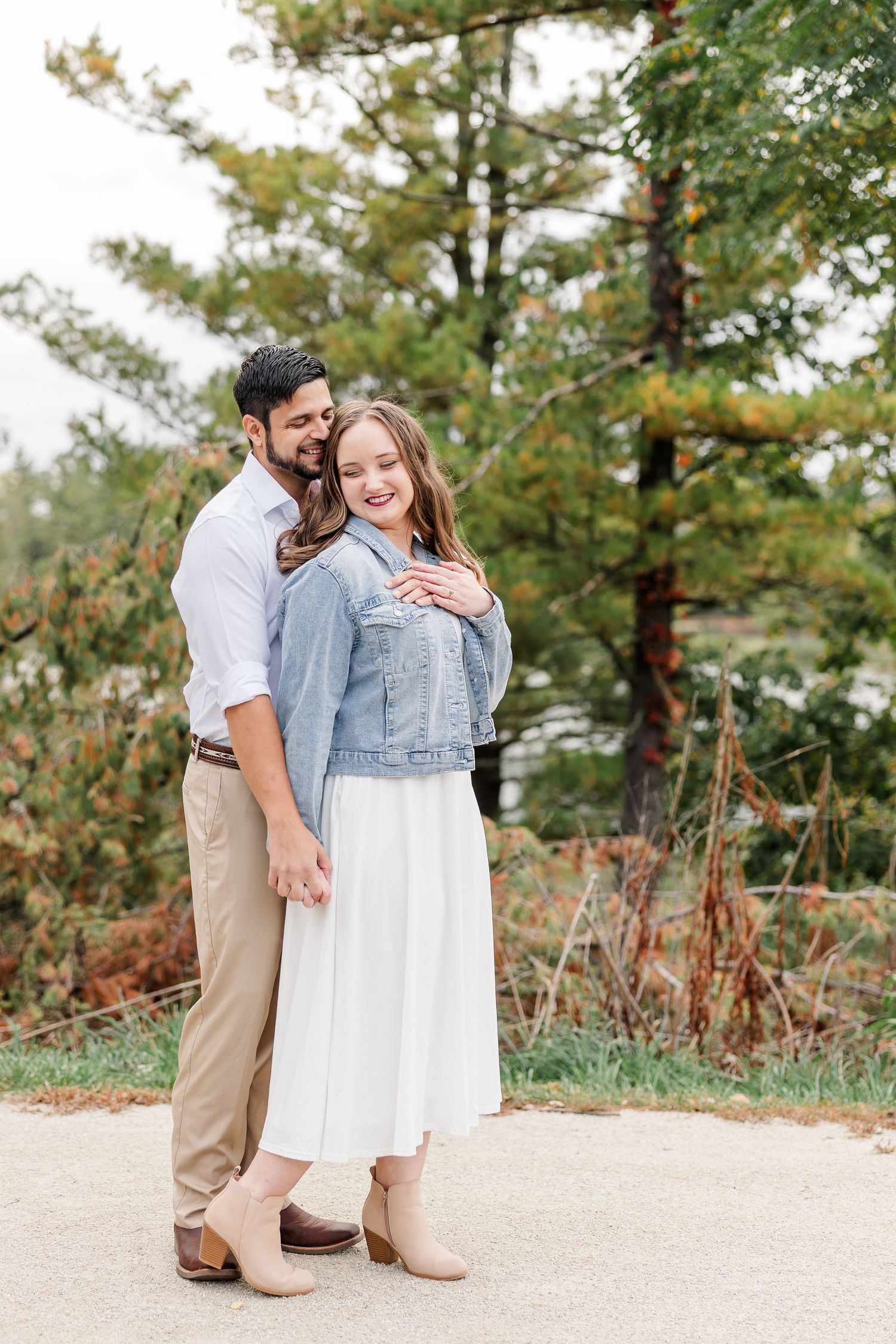 Smiling couple embracing outdoors near pine trees, she wears a denim jacket and white skirt while he hugs her from behind.