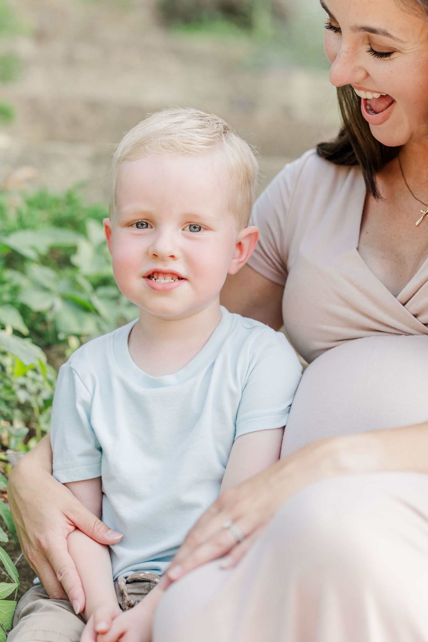 A joyful moment outdoors as a young child sits beside someone in a beige dress among green foliage.