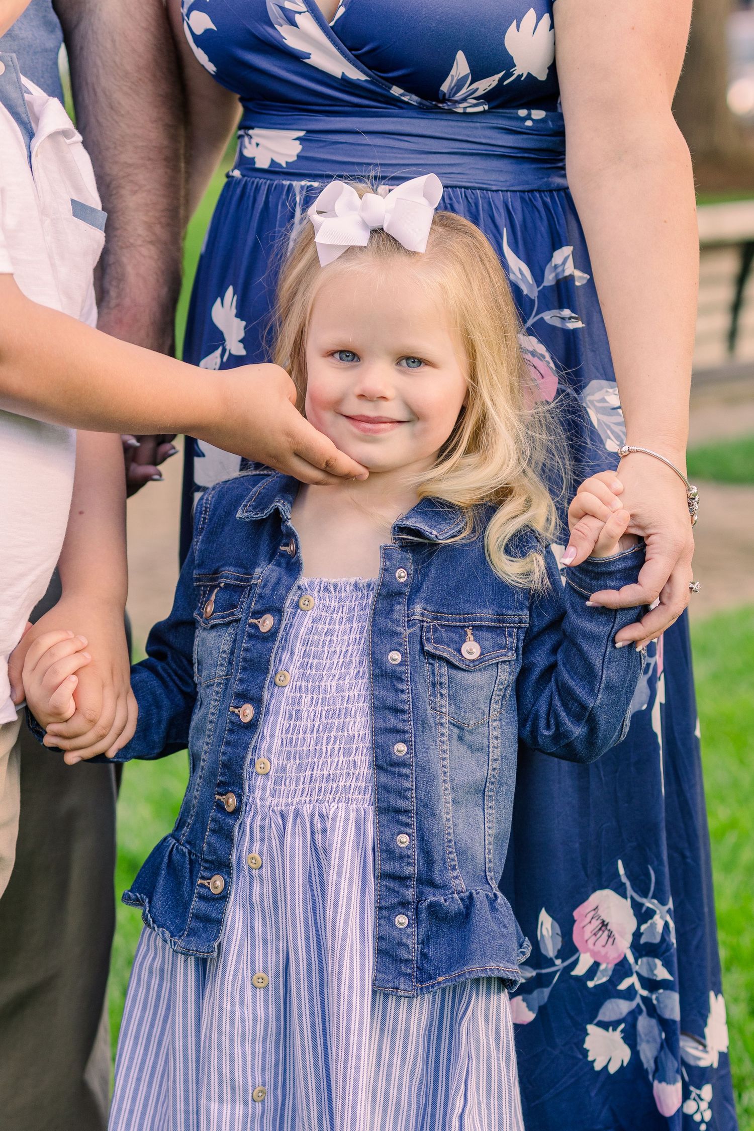 Young girl in denim jacket and striped dress smiles while holding hands with adults.