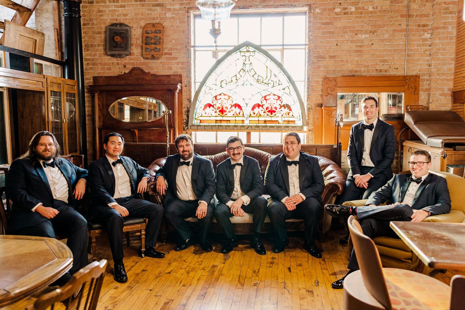 Groomsmen in black tuxedos seated in vintage room with brick walls and arched windows.