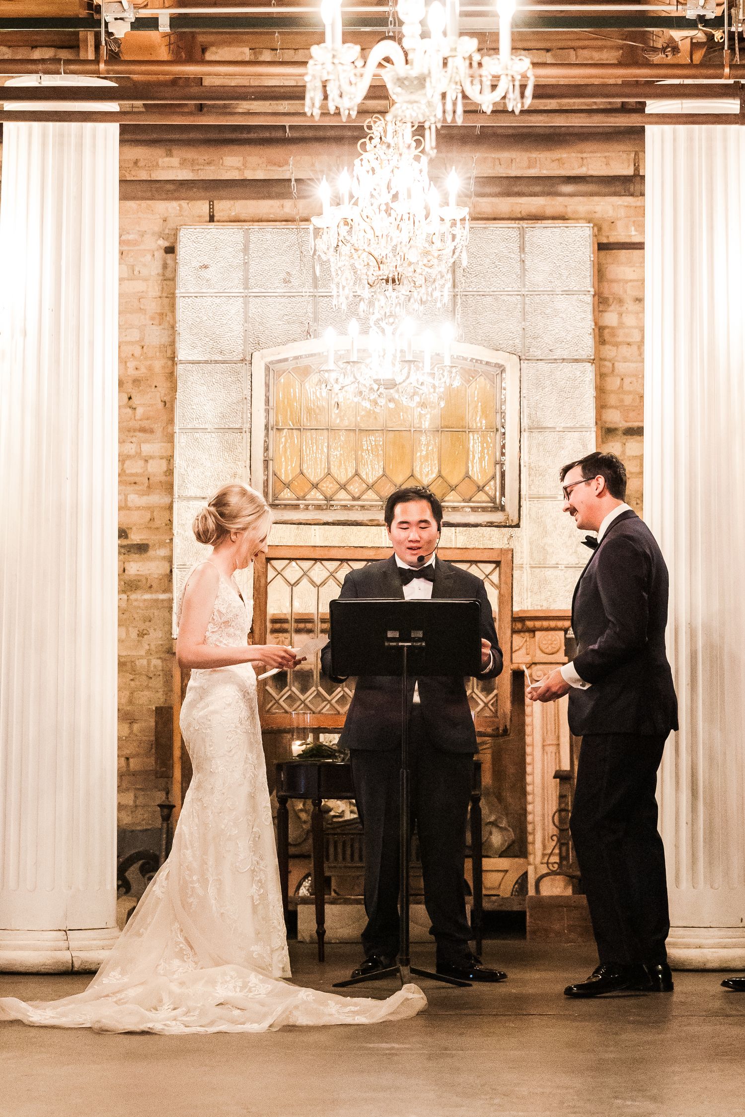 Wedding ceremony under crystal chandelier with brick walls and white columns as backdrop.