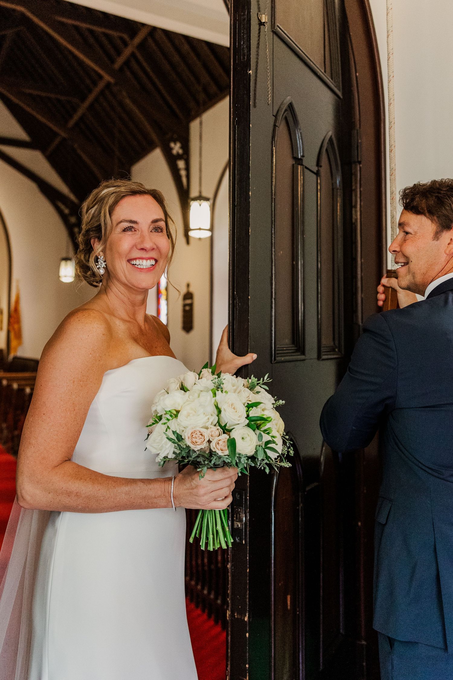 Candid moment of laughter between a couple at church entrance with white bouquet.