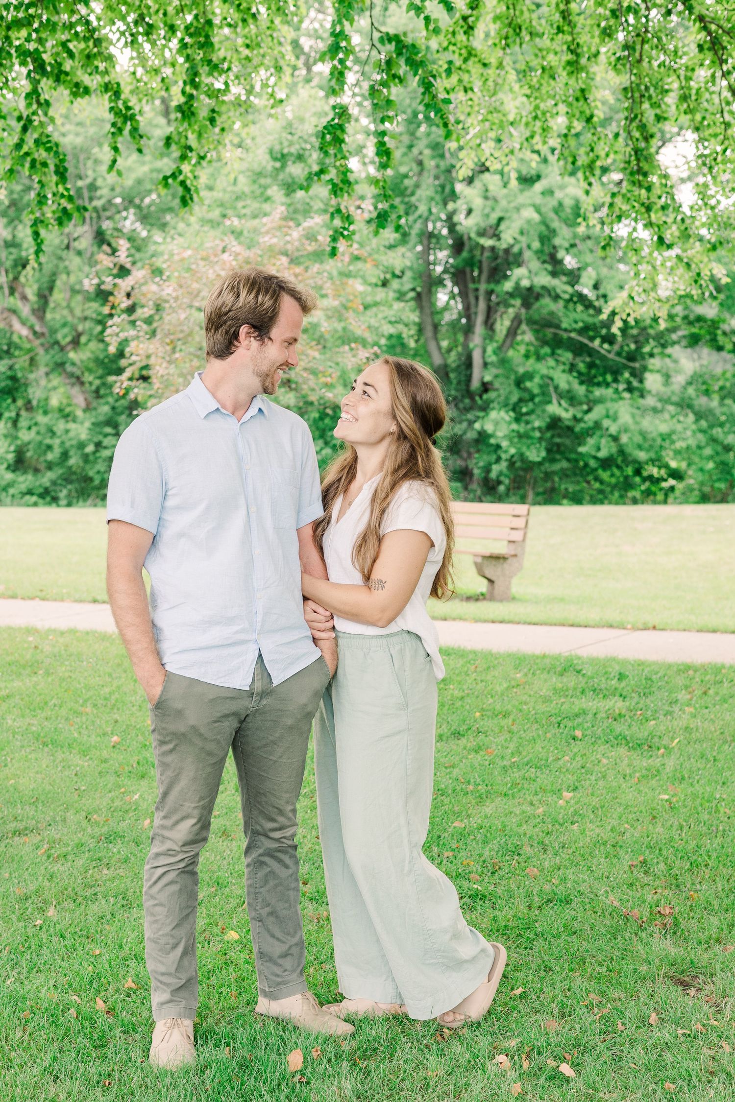 A couple stands together laughing in a scenic park with flowering trees in the background.