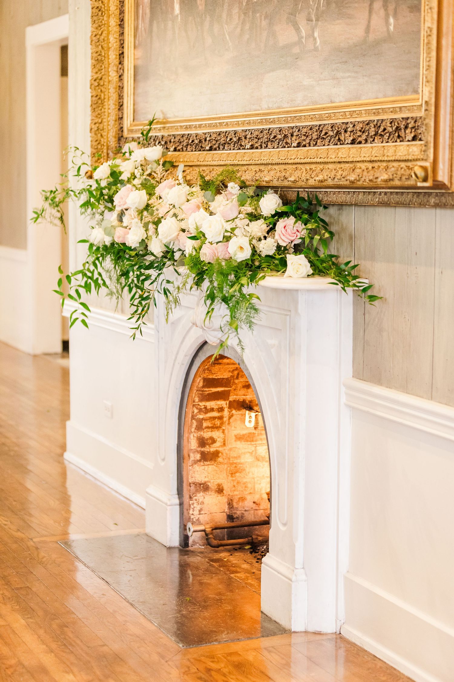 Elegant white fireplace mantel decorated with lush floral arrangements and ornate gold-framed artwork.