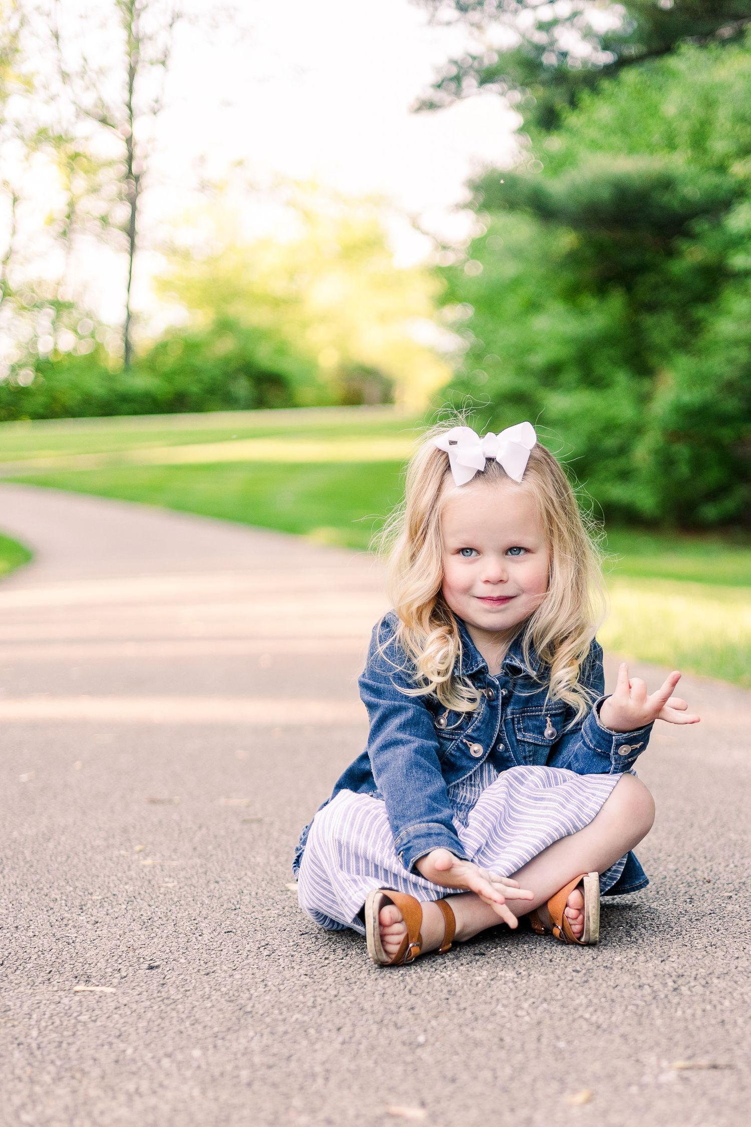 Young child in denim jacket sits cross-legged on a paved path surrounded by green trees.