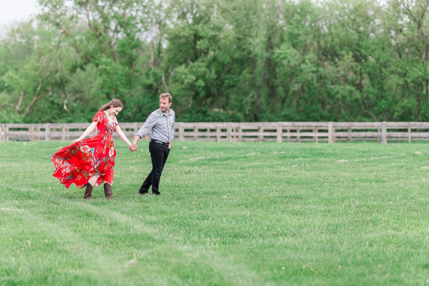 A playful couple runs through a green field with wooden fences in the background.